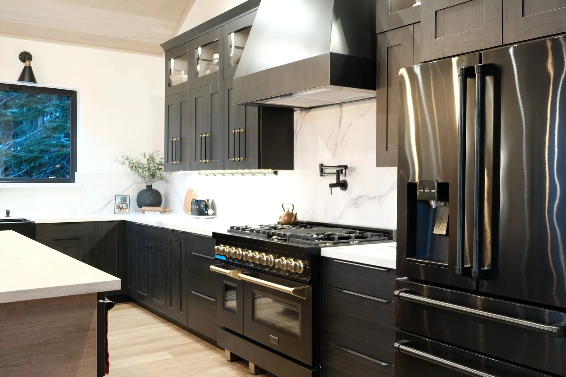 Modern kitchen with dark cabinets, stainless steel refrigerator, gas stove, white marble backsplash, wooden floor, and a window showing outdoor trees.