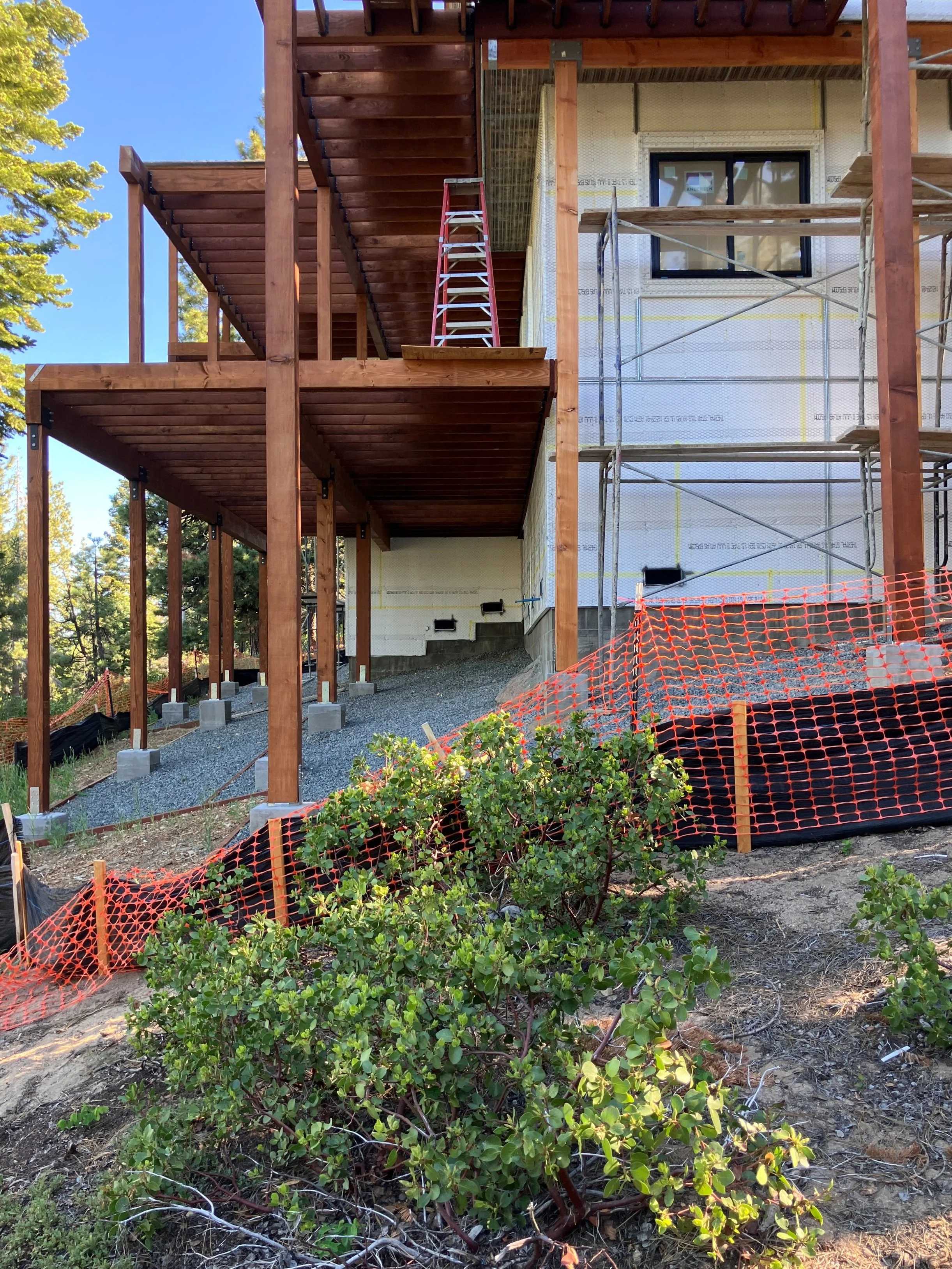 Construction site of a house with wooden decks and scaffolding, orange safety fencing, and a small shrub in the foreground.
