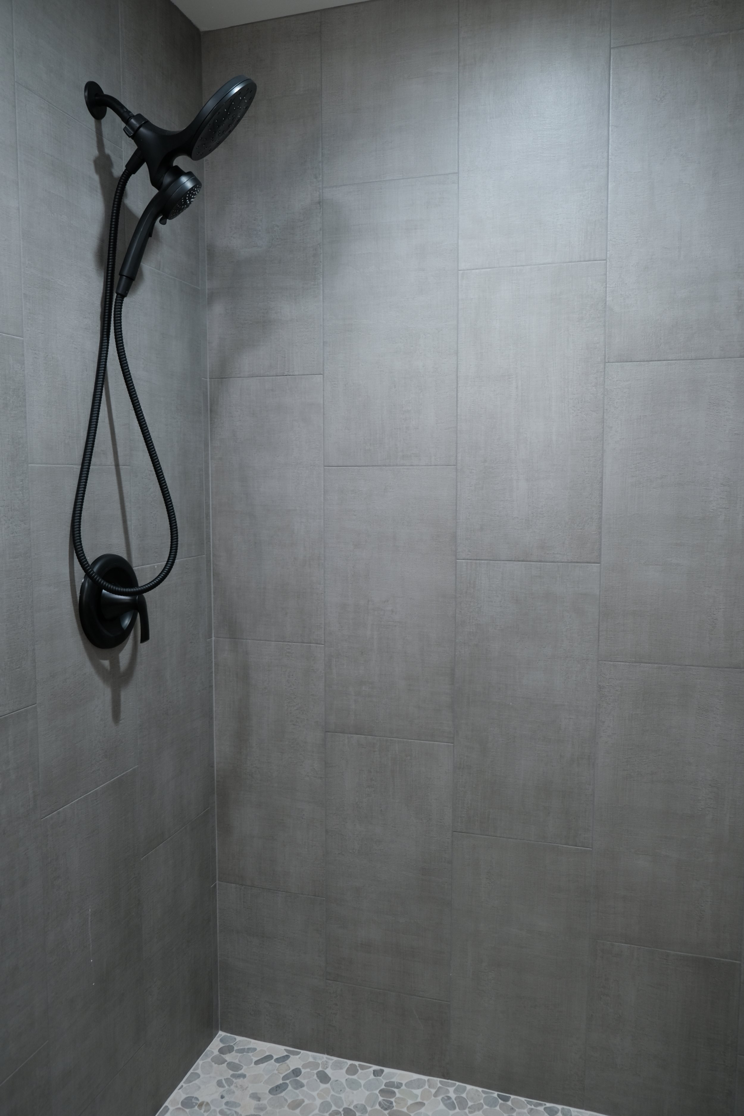 Shower area with gray tiled walls, pebble stone floor, and a black showerhead with a handheld attachment.