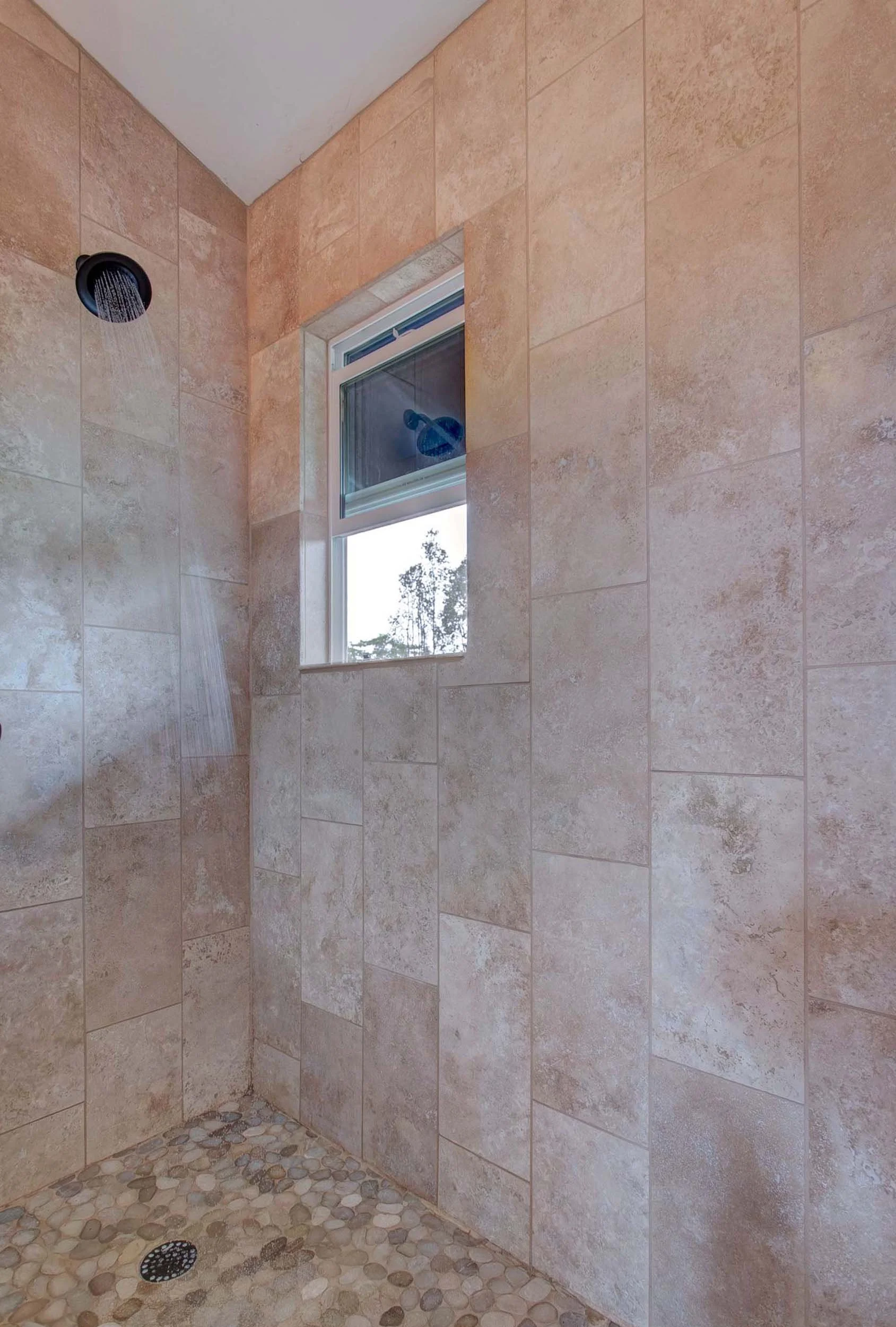 Shower with beige tiled walls, pebble floor, a black shower head, and a small window.