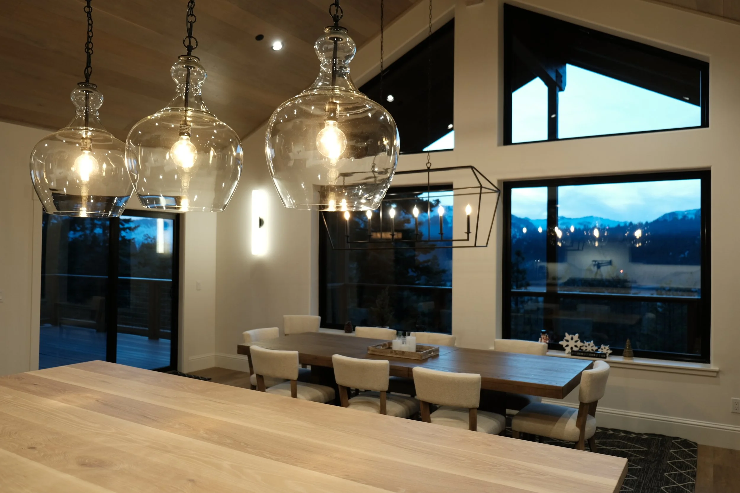 Modern dining room with a wooden table, cream chairs, and hanging glass pendant lights, overlooking large windows with mountain views.