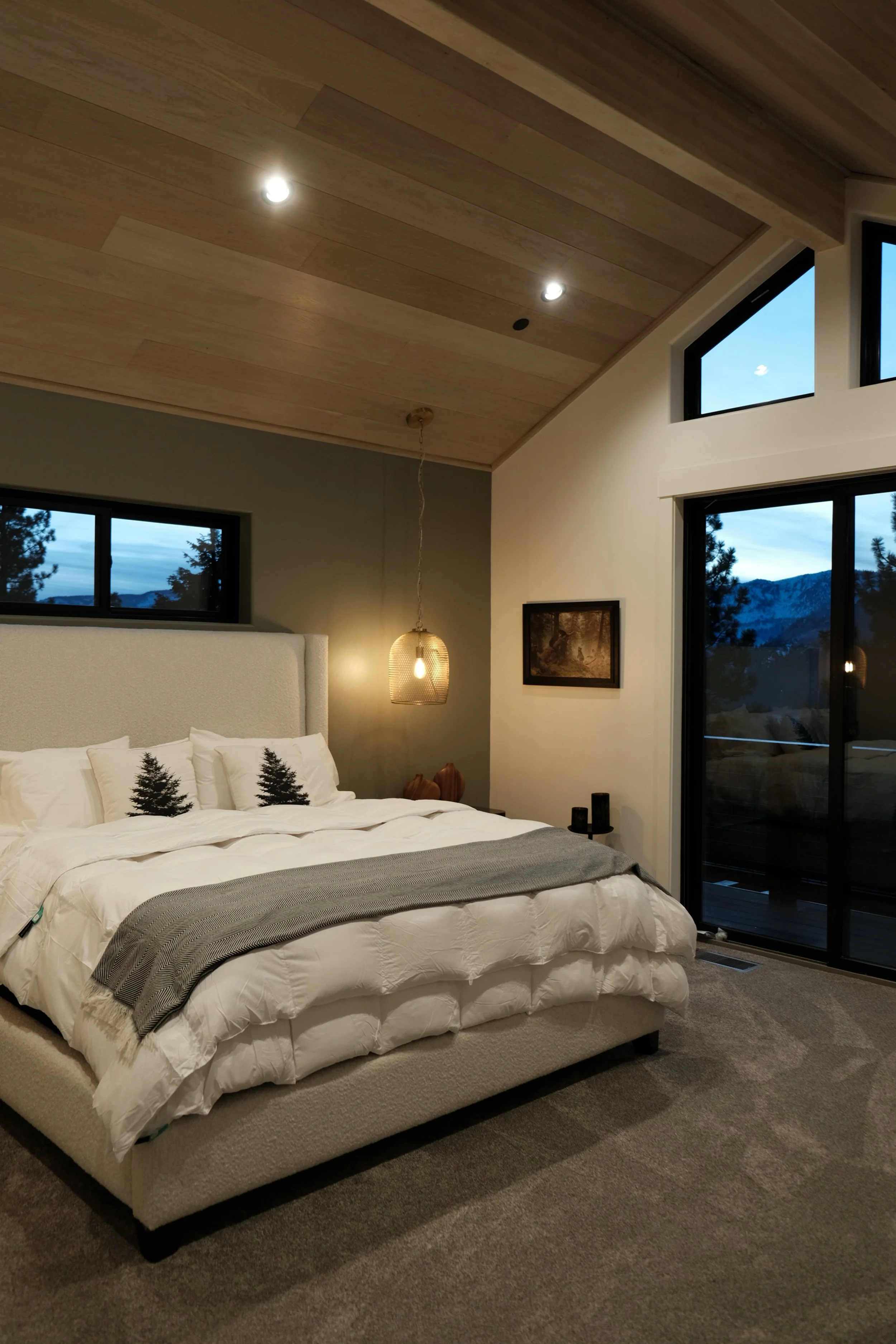 Modern bedroom with vaulted wooden ceiling, large windows showing mountain views, white bedding with tree-themed pillows, and warm ambient lighting.