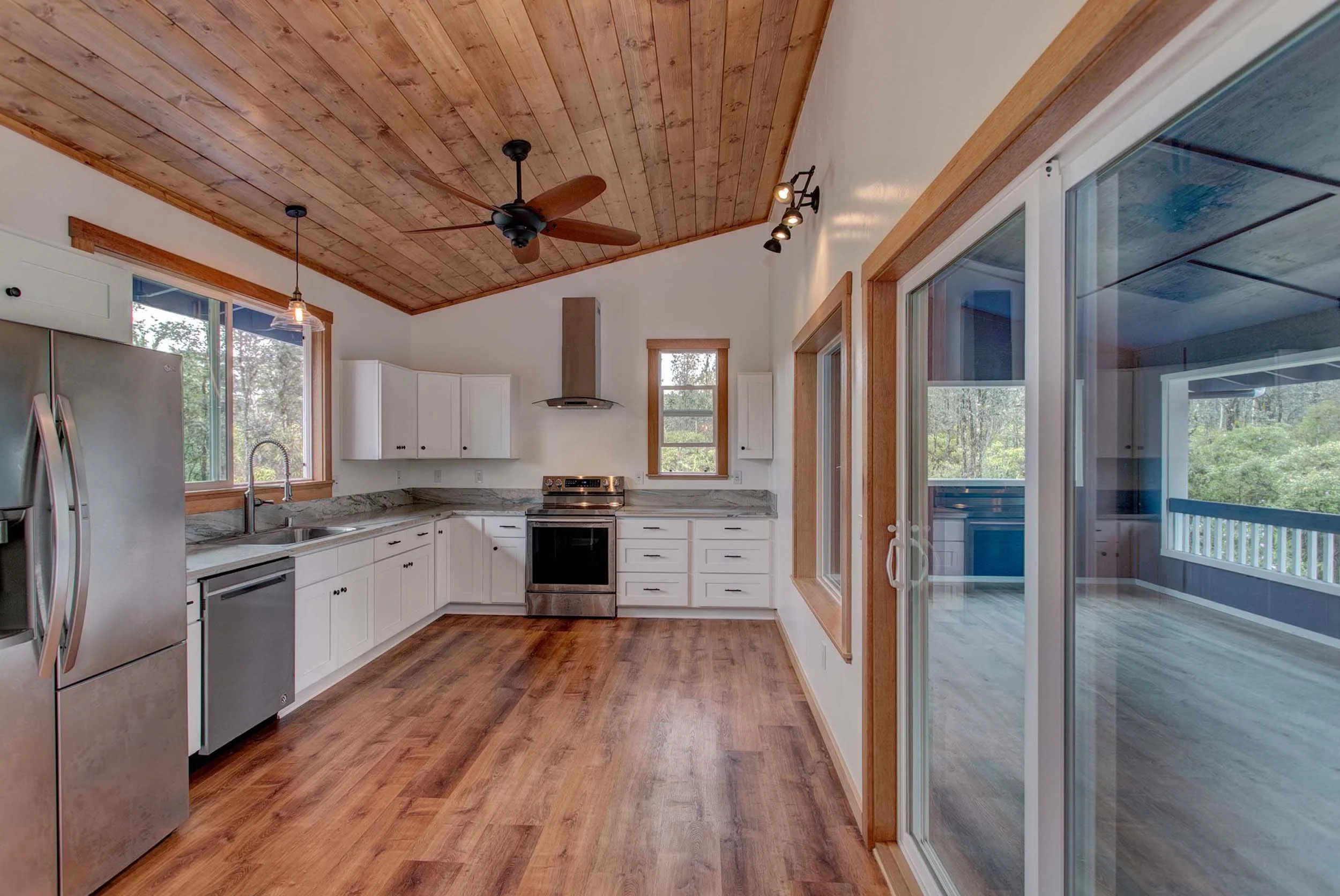 Kitchen with white cabinets, stainless steel appliances, wooden floor, and a wood-paneled ceiling. There is a sliding glass door leading to an outdoor deck.