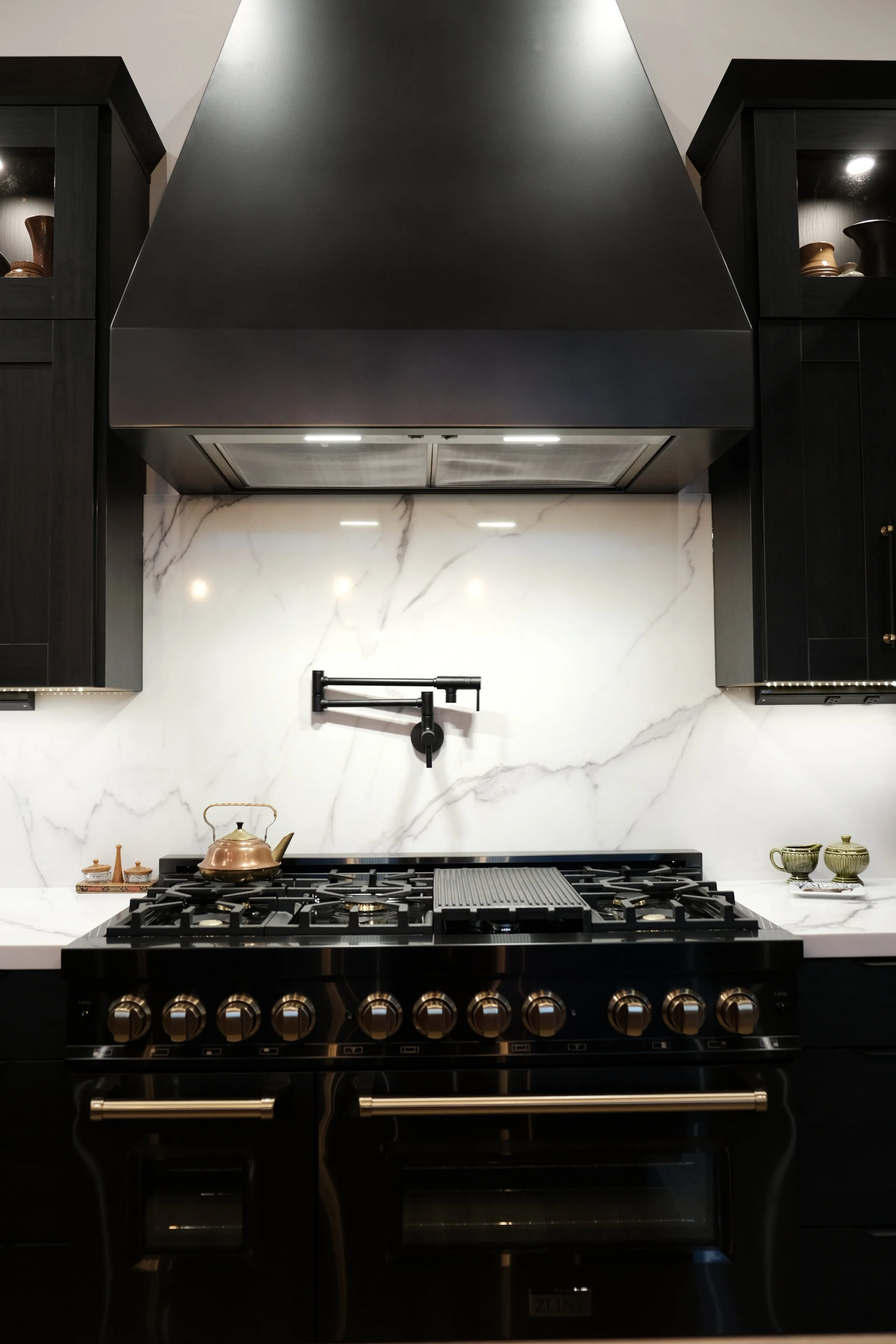 Modern kitchen with black cabinetry, marble backsplash, and a black gas stove with brass knobs underneath a large black range hood.