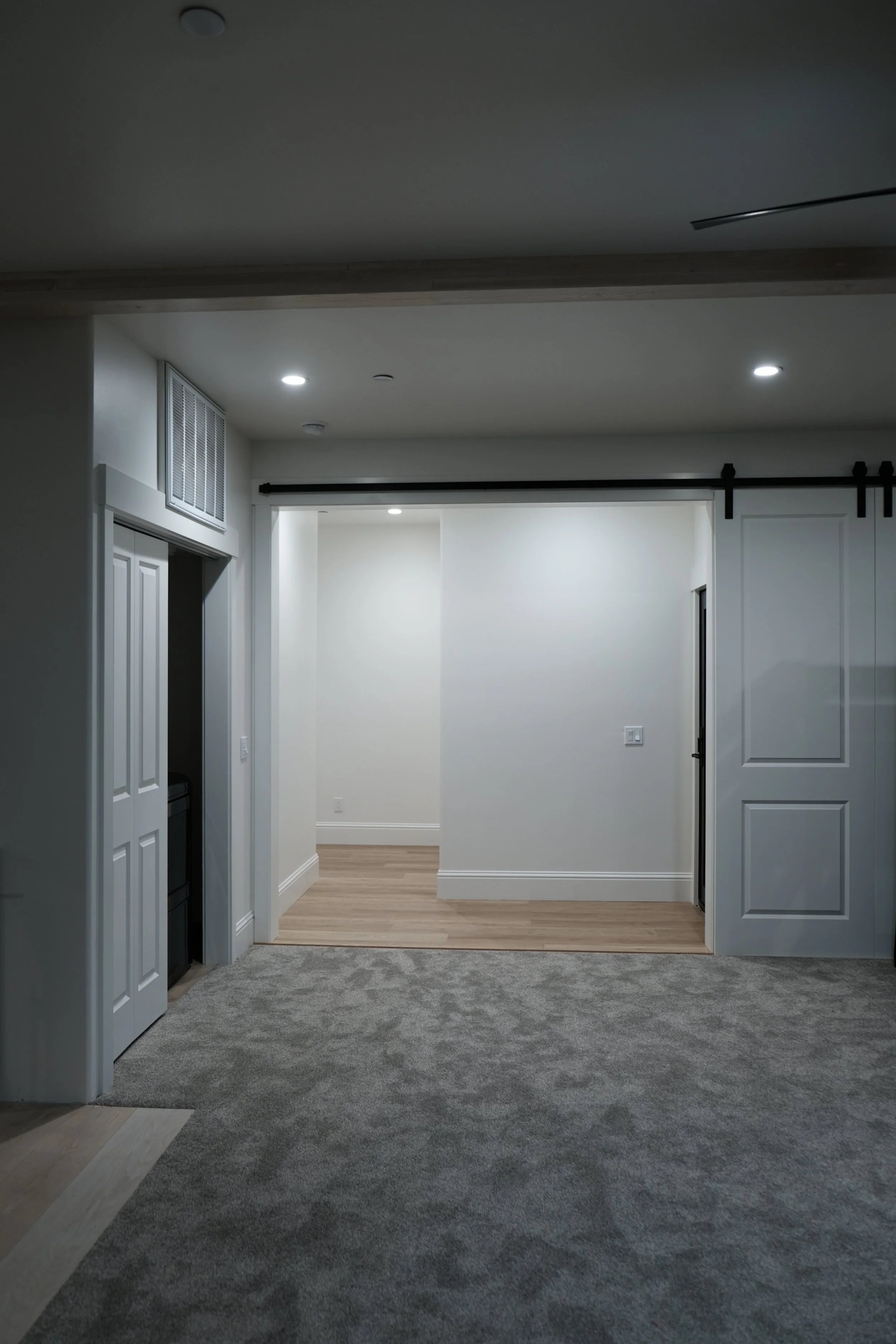 Empty modern living room with gray carpet, white walls, and openings to hardwood floors, ceiling lights, and sliding barn door.