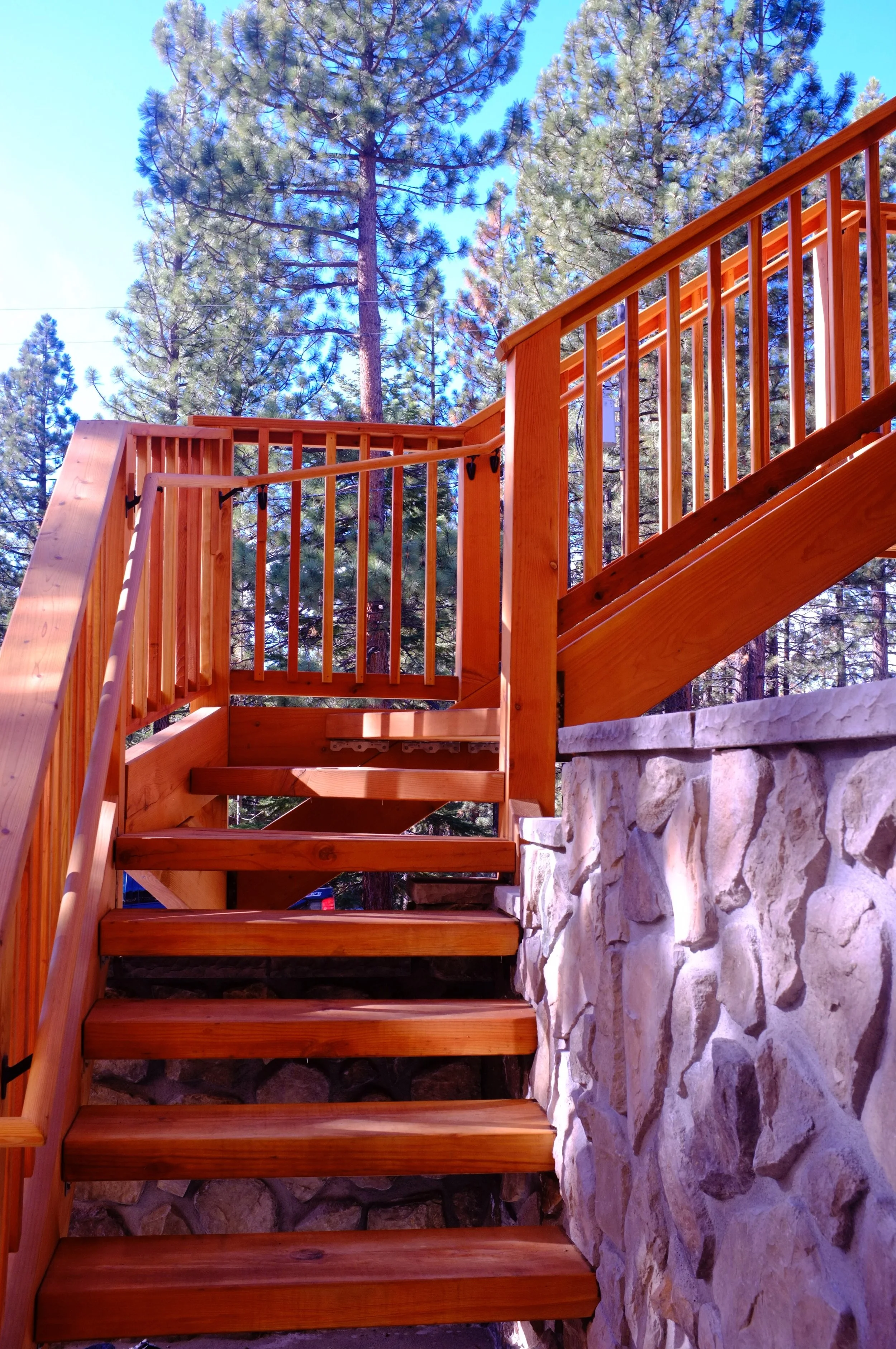 Wooden outdoor staircase with railing leading up to a deck, stone foundation at the base, surrounded by tall pine trees, under a blue sky.