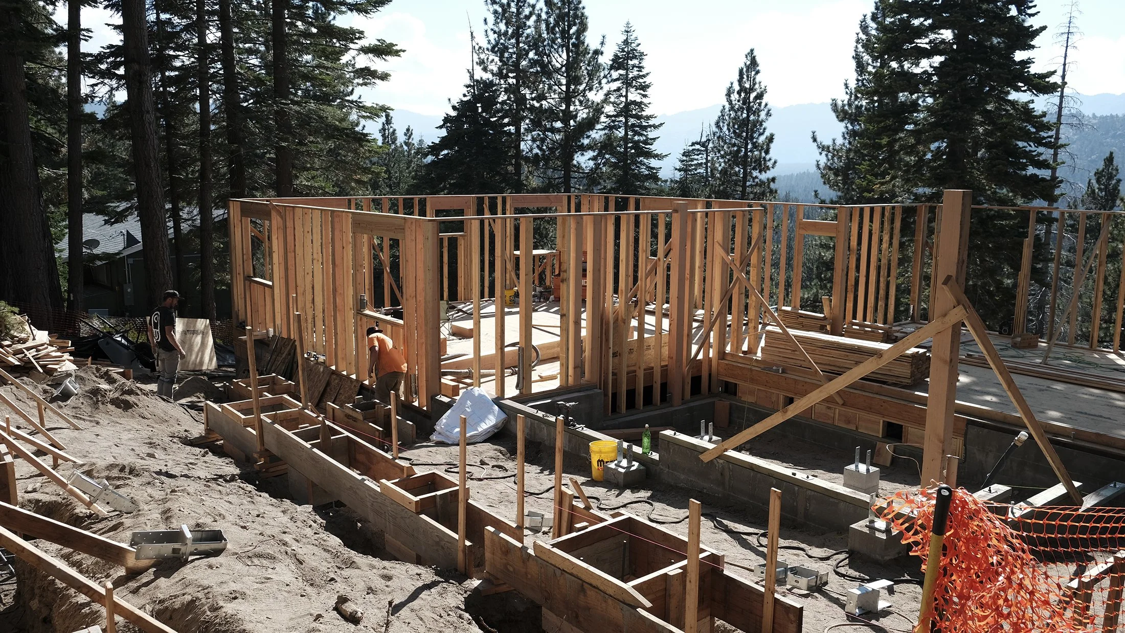 Building construction site with wooden framing in a forested area, two workers installing wood beams, surrounded by construction materials and equipment.