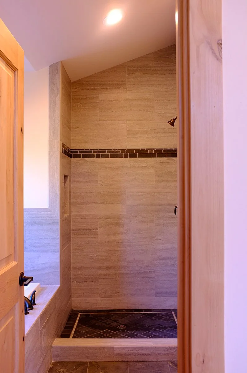 A shower area with beige tiles, a horizontal strip of small dark tiles, and a bronze shower head. The shower has a dark tiled floor and is partially visible through a doorway with a wooden door frame.