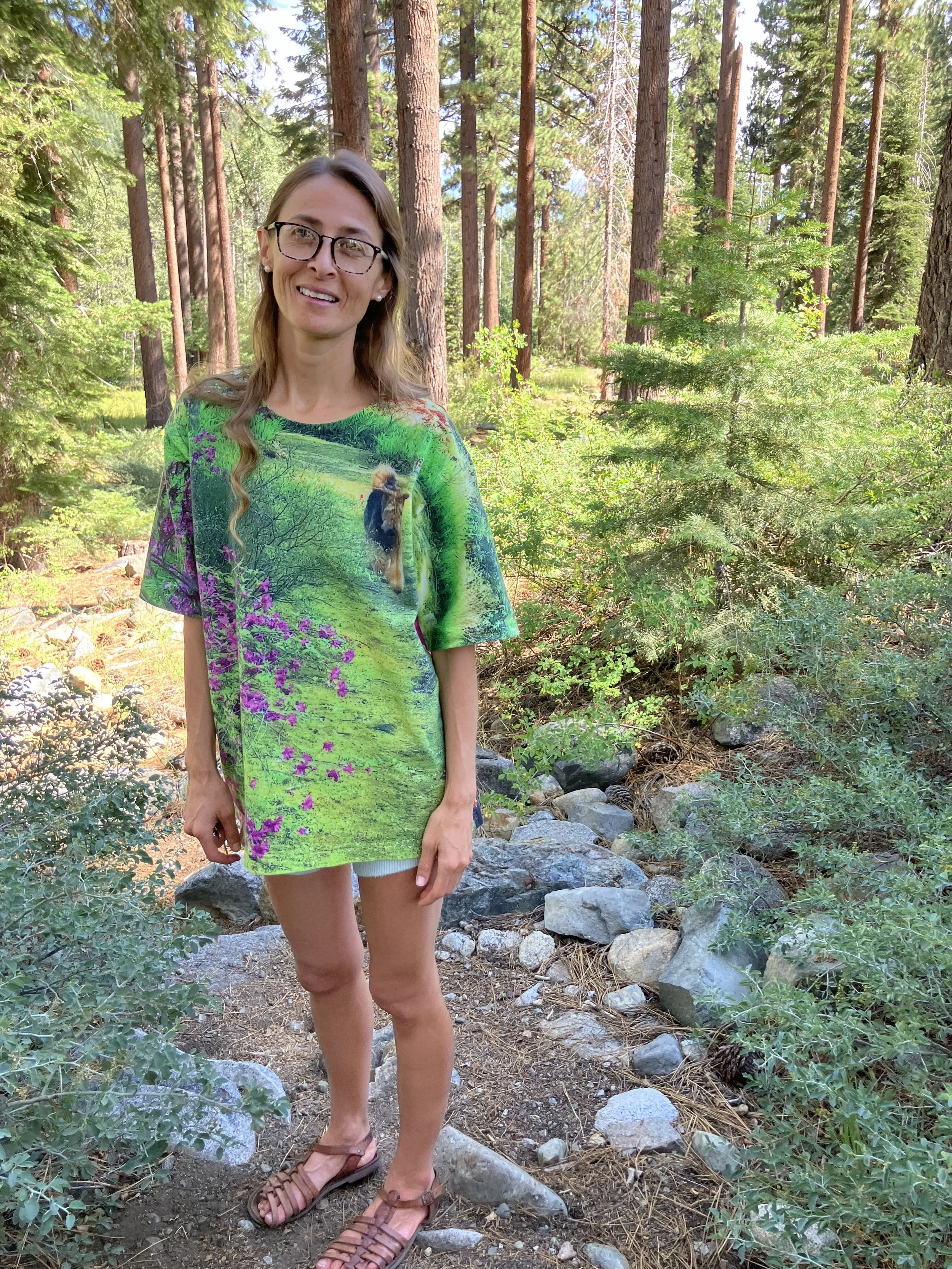 A woman with glasses and long hair standing on a rocky forest trail surrounded by pine trees and greenery, smiling at the camera.
