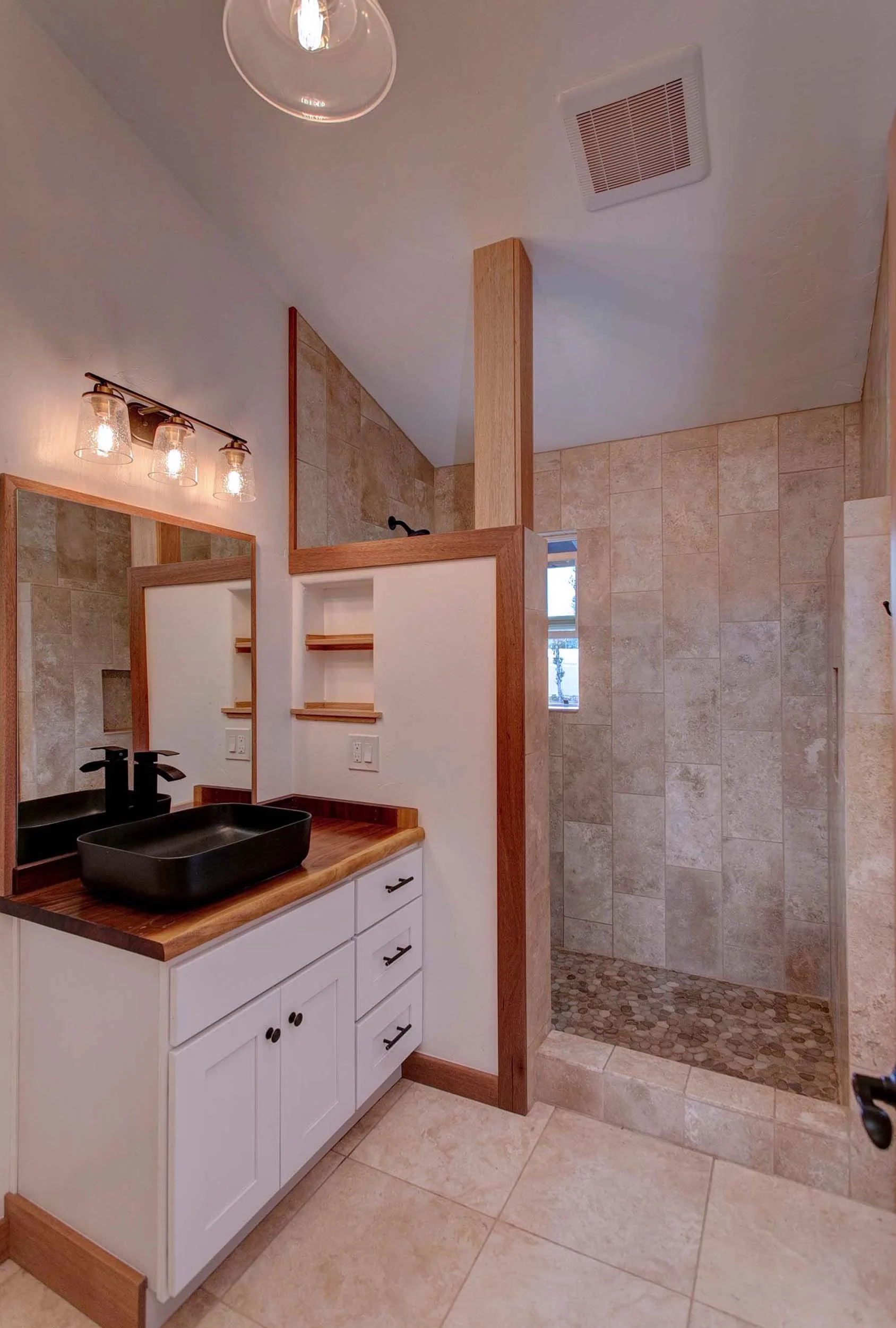 Bathroom with a white vanity, black vessel sink, black faucet, mirror, wall-mounted light fixtures, and a walk-in shower with beige tiles and pebble floor.