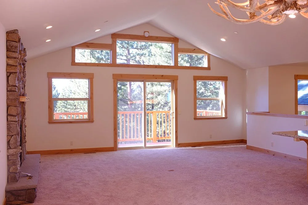 Empty living room with large windows, sliding glass door leading to an outdoor balcony, wooden trim, and a stone fireplace on the left.