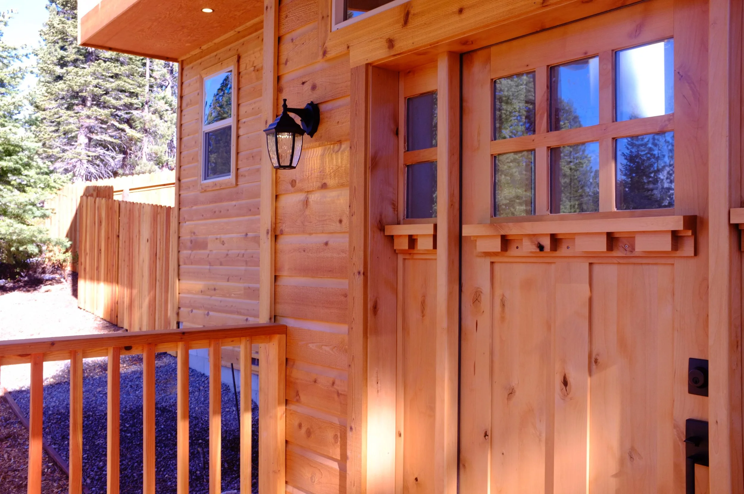 Close-up view of a wooden house exterior with a door, window, outdoor light fixture, wooden fence, and trees in the background.