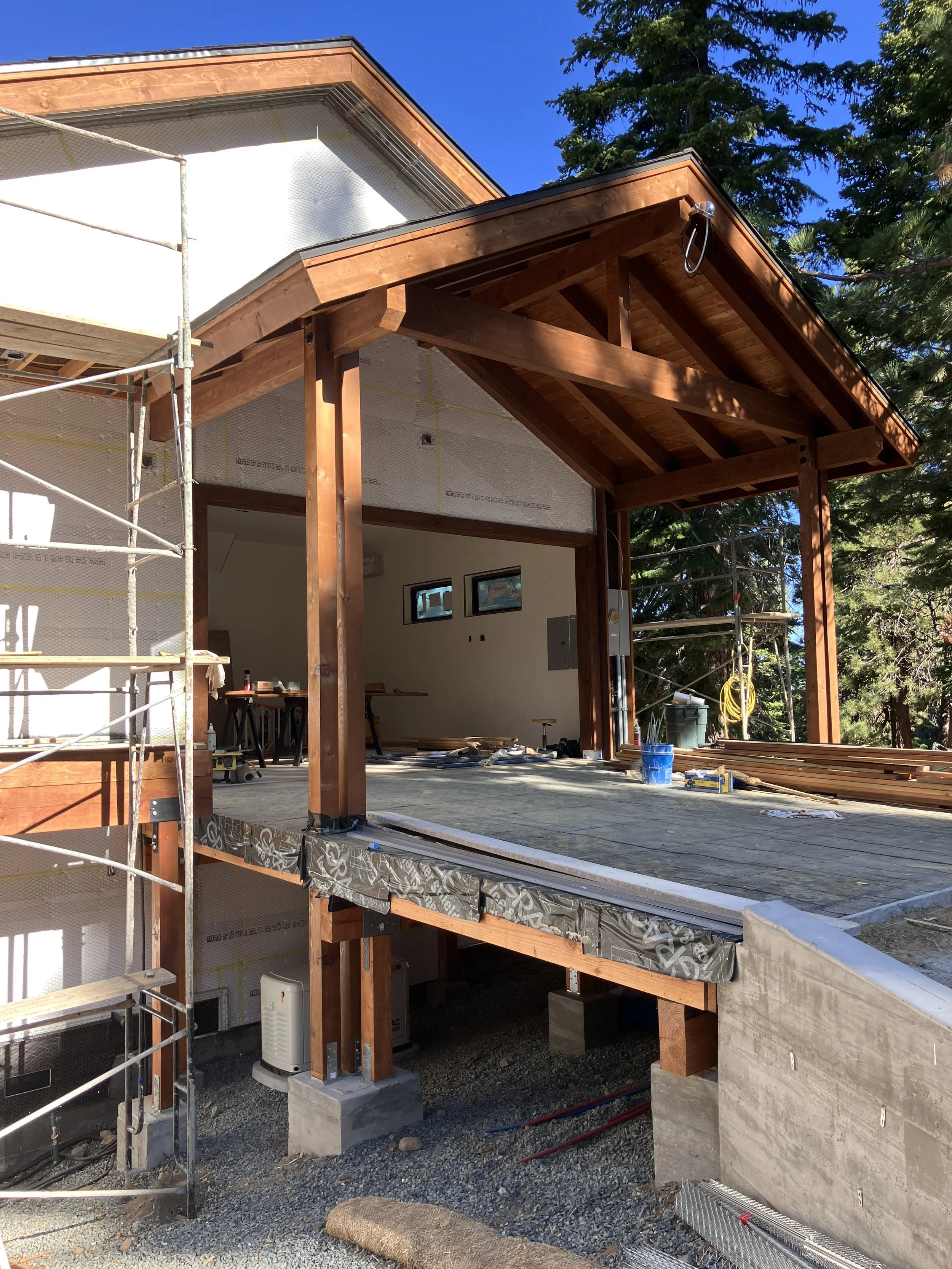 Construction site of a residential building with wooden framing, porch, and scaffolding, showing ongoing work and building materials.