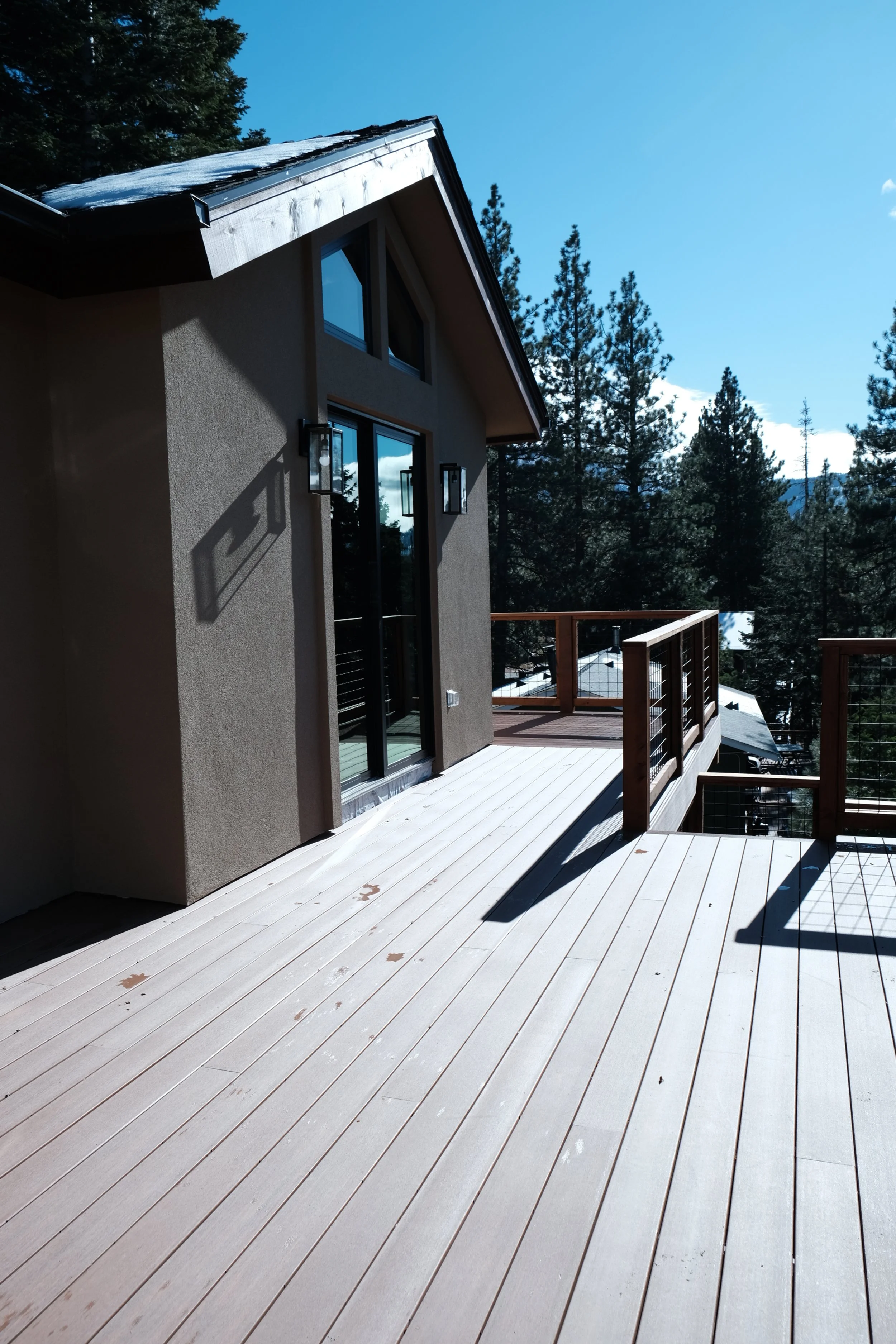 A wooden deck outside a house with glass sliding doors, surrounded by a forest of tall pine trees on a clear day.