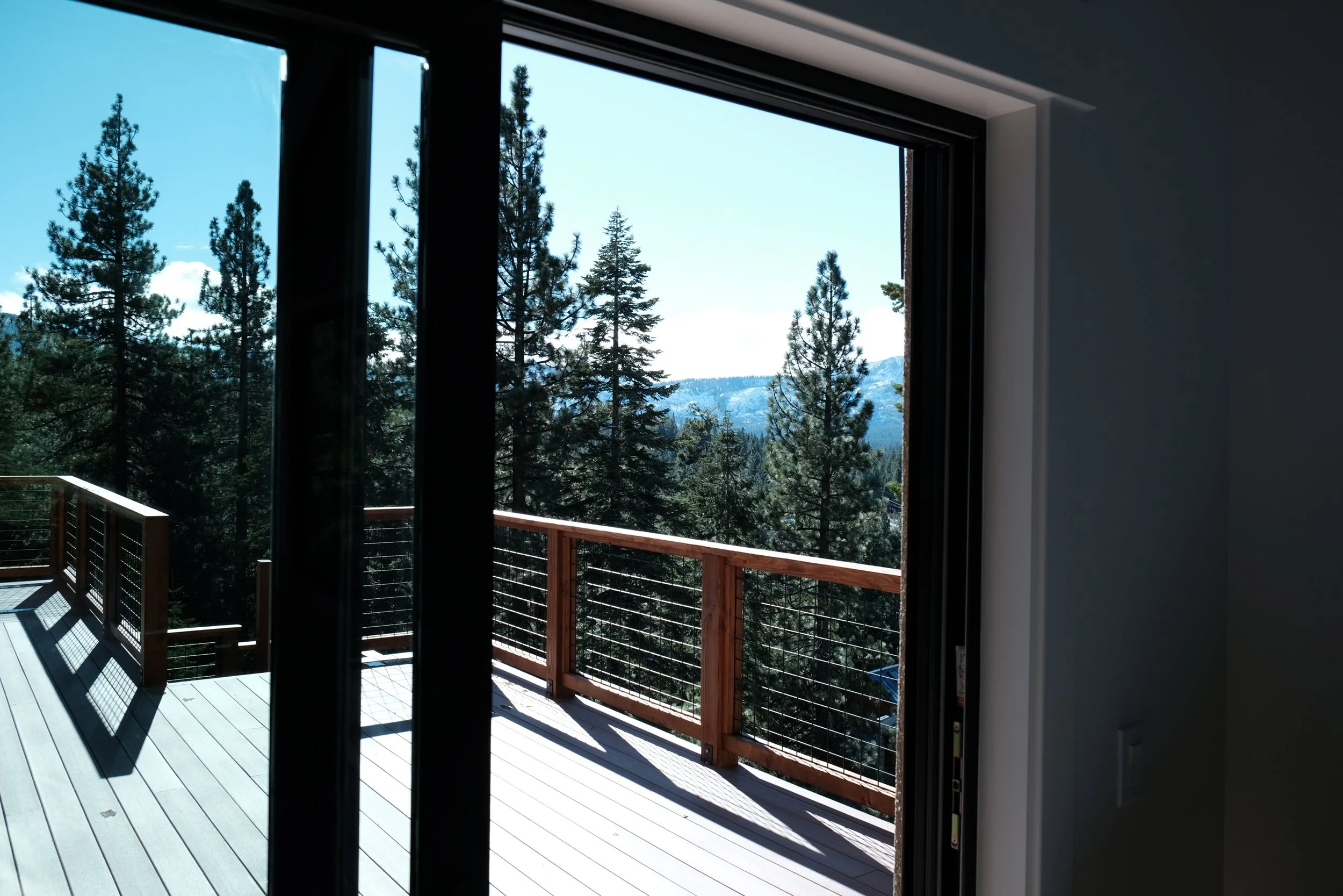 View of a wooden balcony overlooking a forest of pine trees with mountains in the distance on a sunny day.