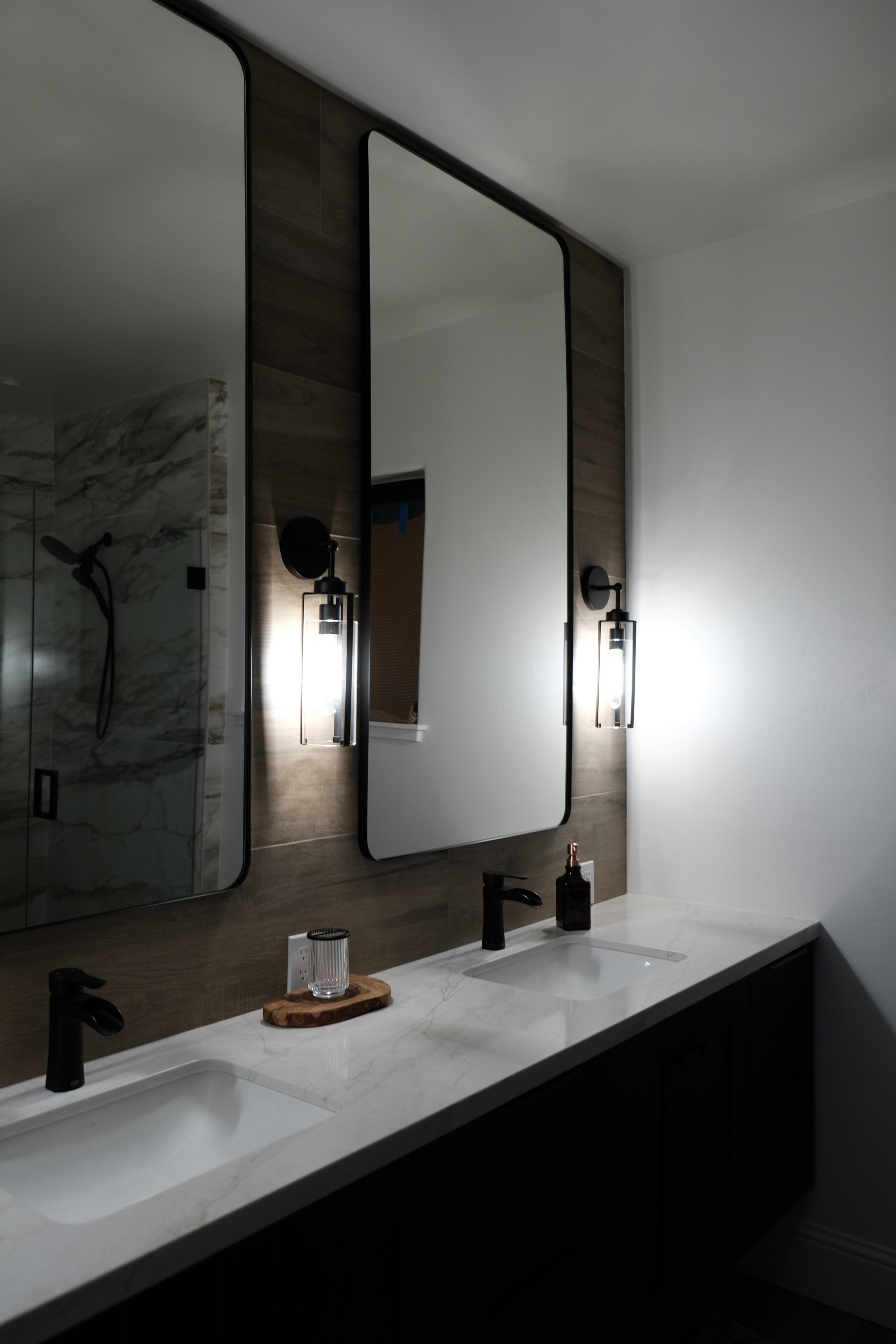 Modern bathroom with a double vanity, marble countertop, black fixtures, large mirror, and wall-mounted lights. Shower area with glass door and marble wall is visible in the background.