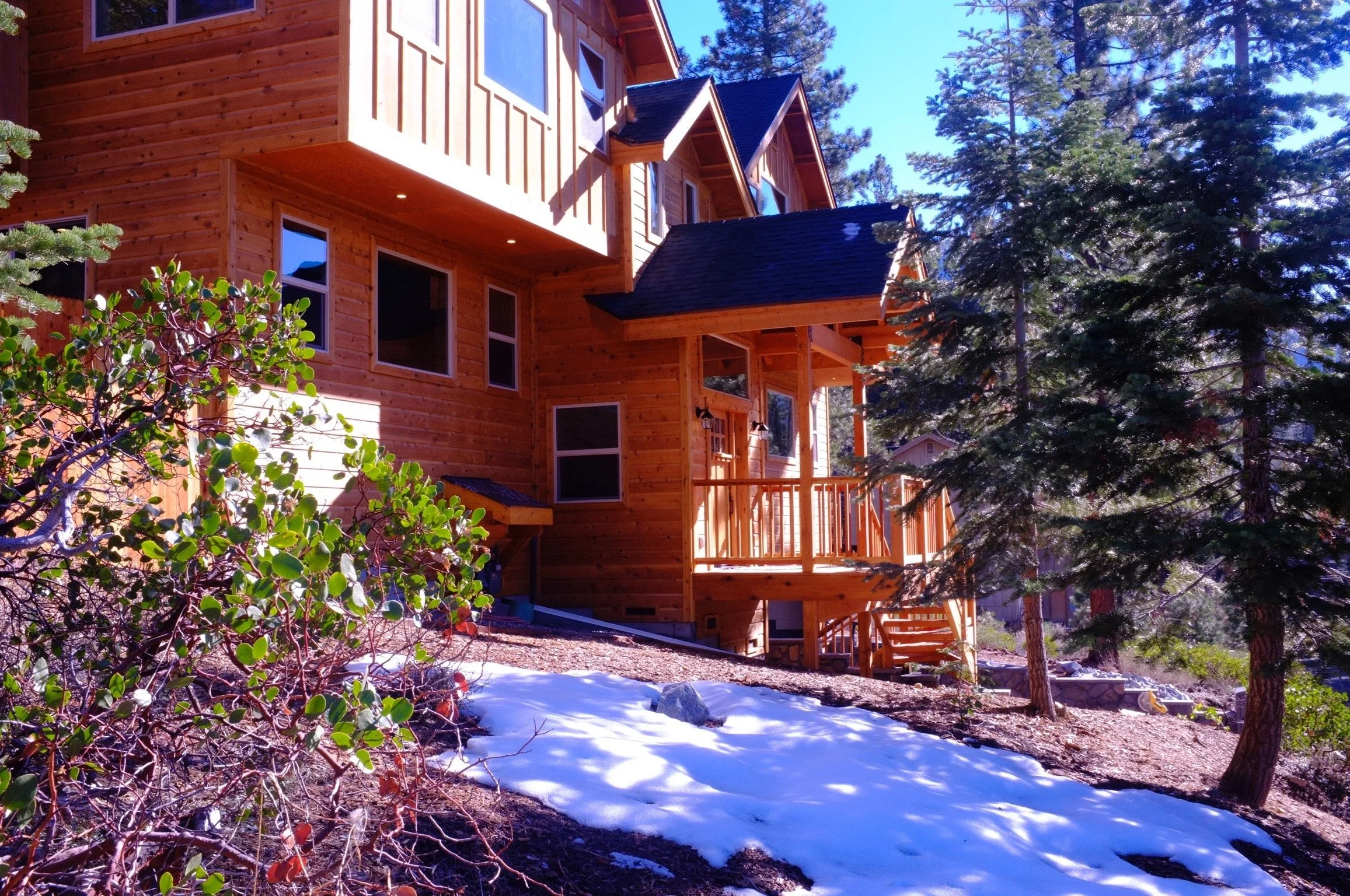 A wooden house in a forested area with snow on the ground, tall evergreen trees, and a bright blue sky.