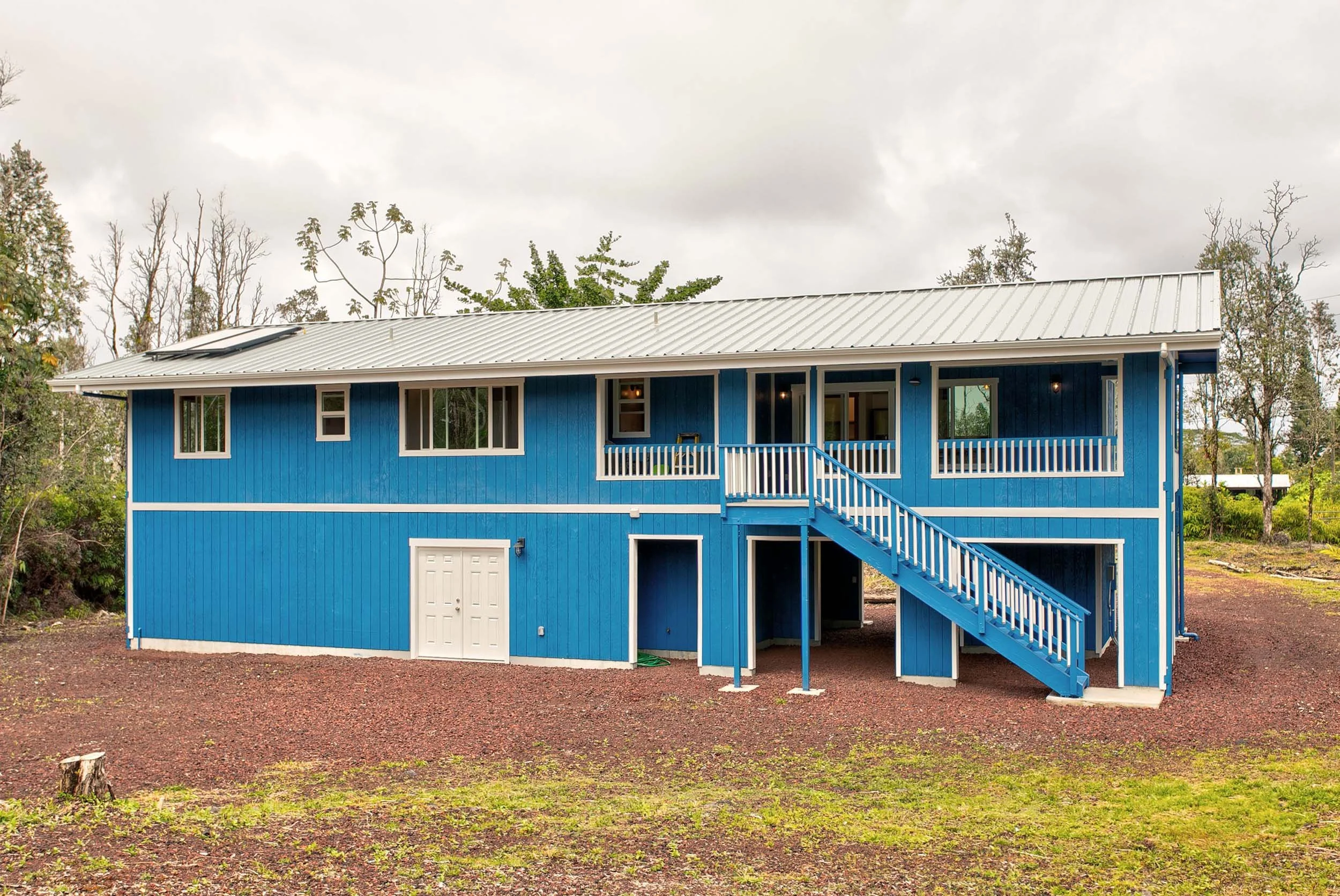 Blue house with white door, staircase, and railings, built on a gravel foundation, surrounded by grass and trees under an overcast sky.