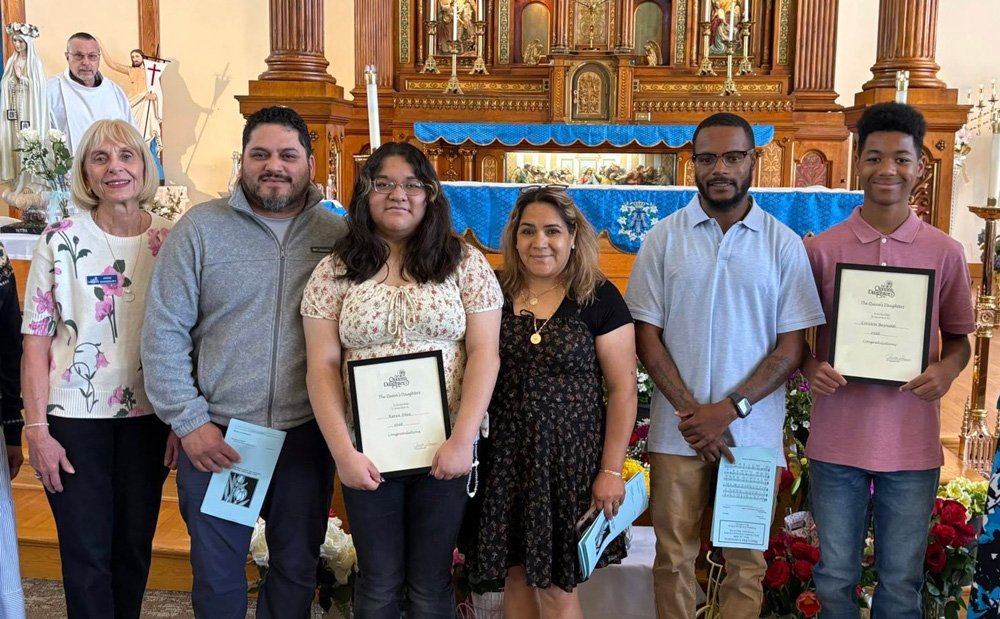 Group of six people posing inside a church, three of whom are holding framed certificates, with religious statues and an altar in the background.