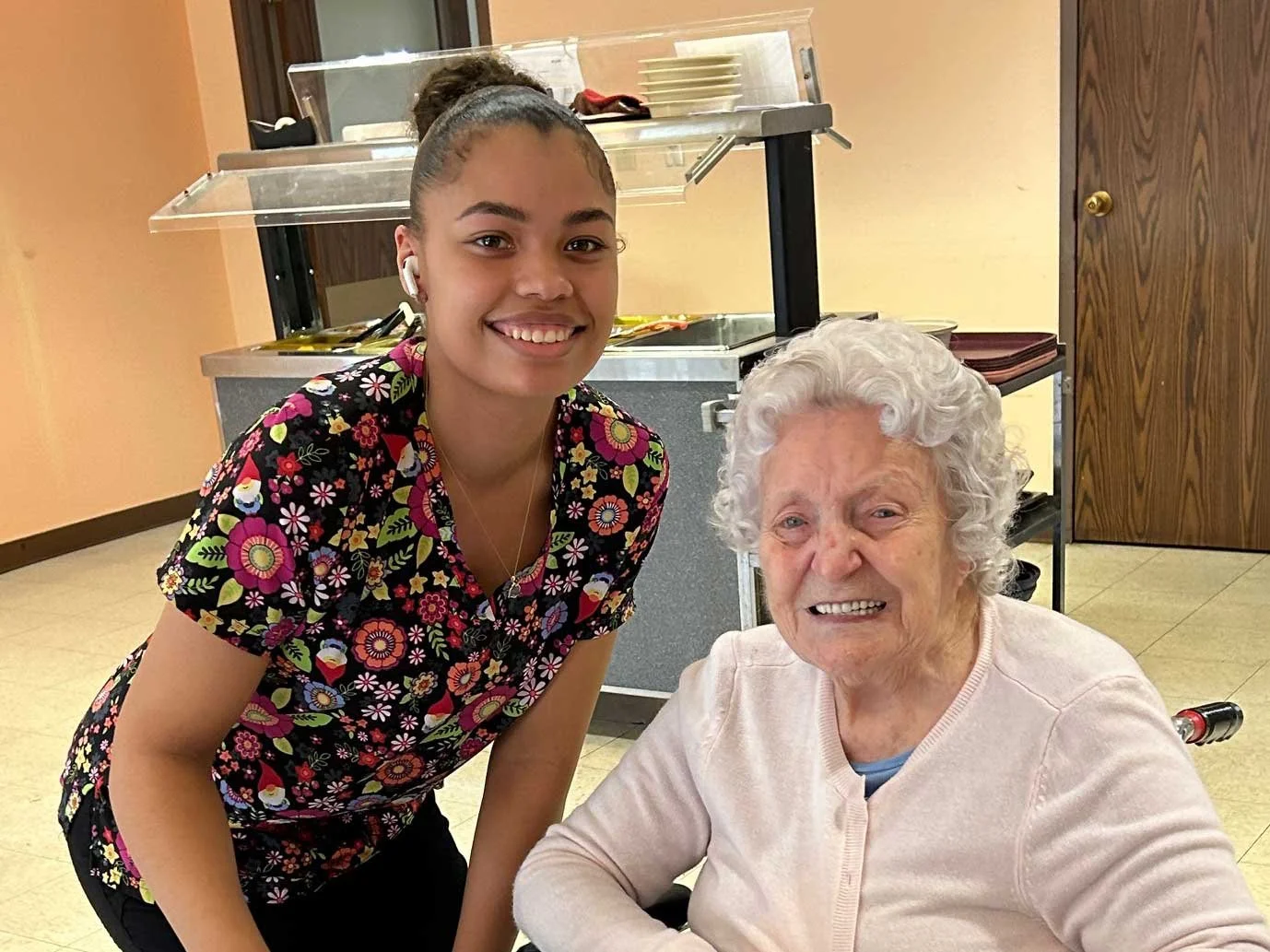 A young woman and an elderly woman smile for a photo in what appears to be a cafeteria or dining area, with a food service counter in the background.