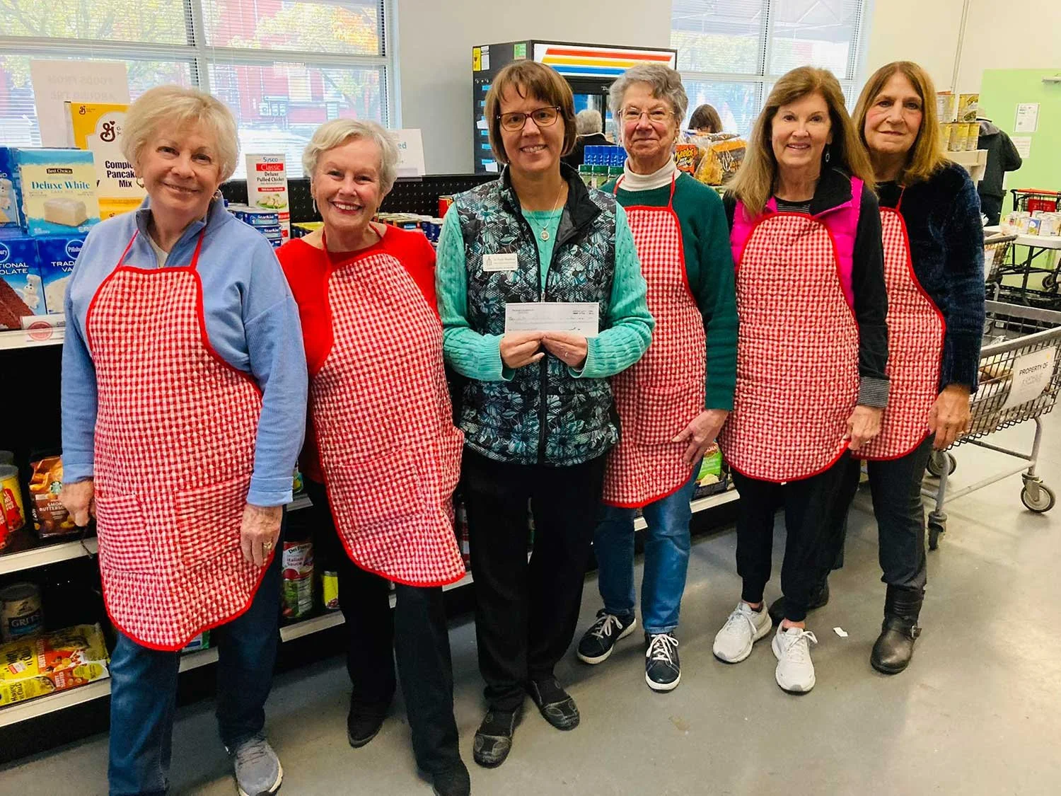 Six women standing in a grocery store, wearing red and white checkered aprons. One woman in the middle is holding a check, and they are all smiling. There are shelves of grocery items behind them.