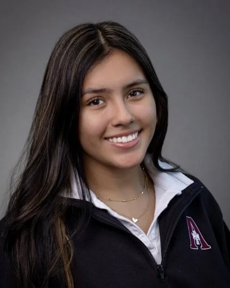 Portrait of a young woman with long dark hair, smiling, wearing a white collared shirt and a black zip-up jacket with a purple logo, against a gray background.