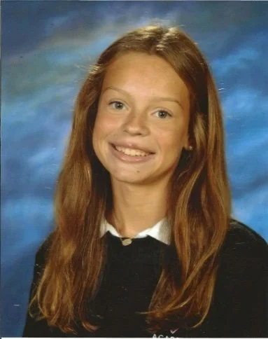 A young girl with long, wavy red hair and a bright smile, wearing a black sweater over a white collared shirt, in front of a blue and green background.