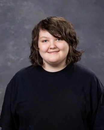 A young woman with curly brown hair wearing a black shirt, smiling against a gray background.