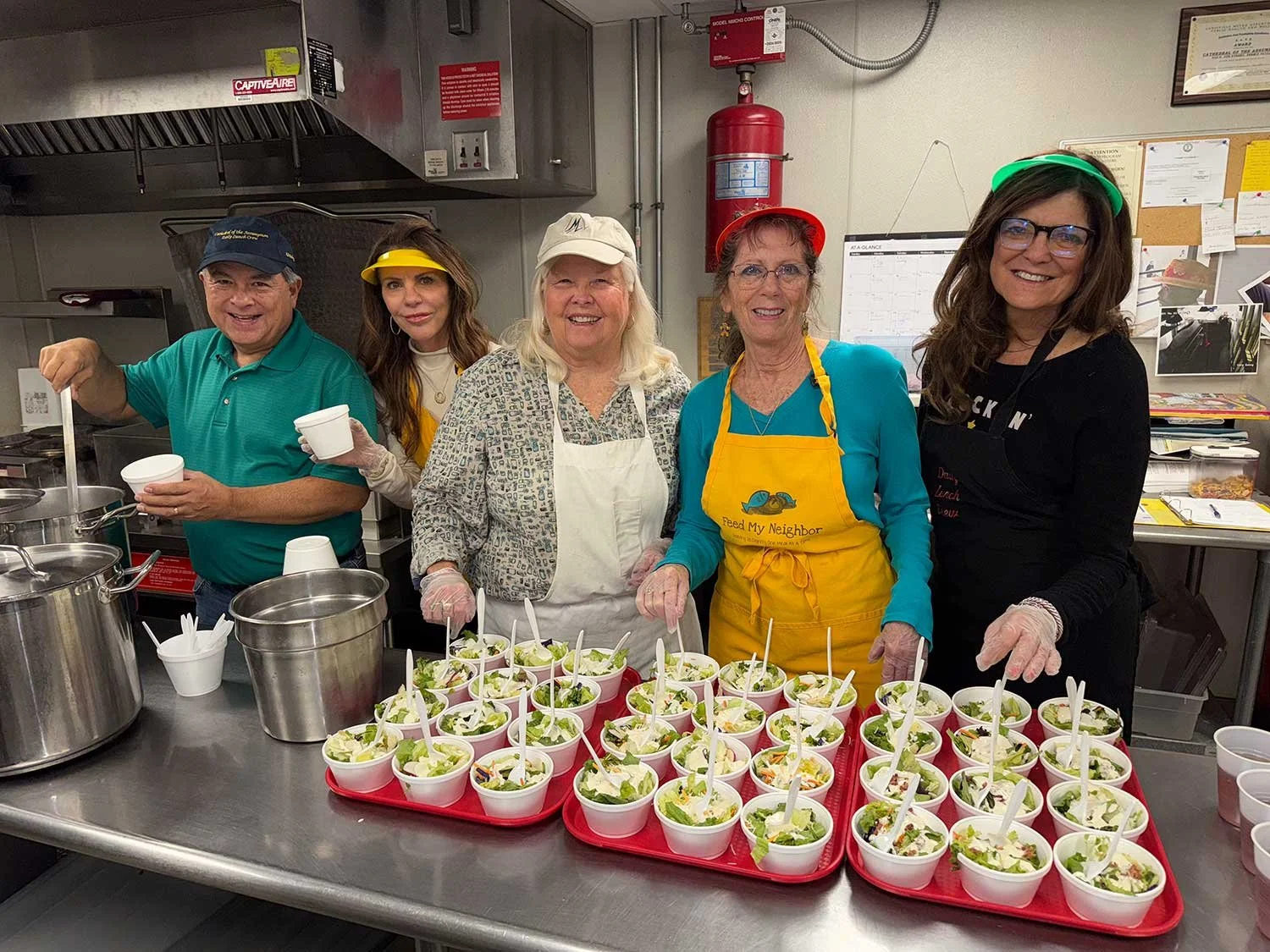 Five people standing behind a table filled with bowls of salad, preparing food together in a kitchen. They are wearing aprons, gloves, and hair coverings, and are smiling at the camera.