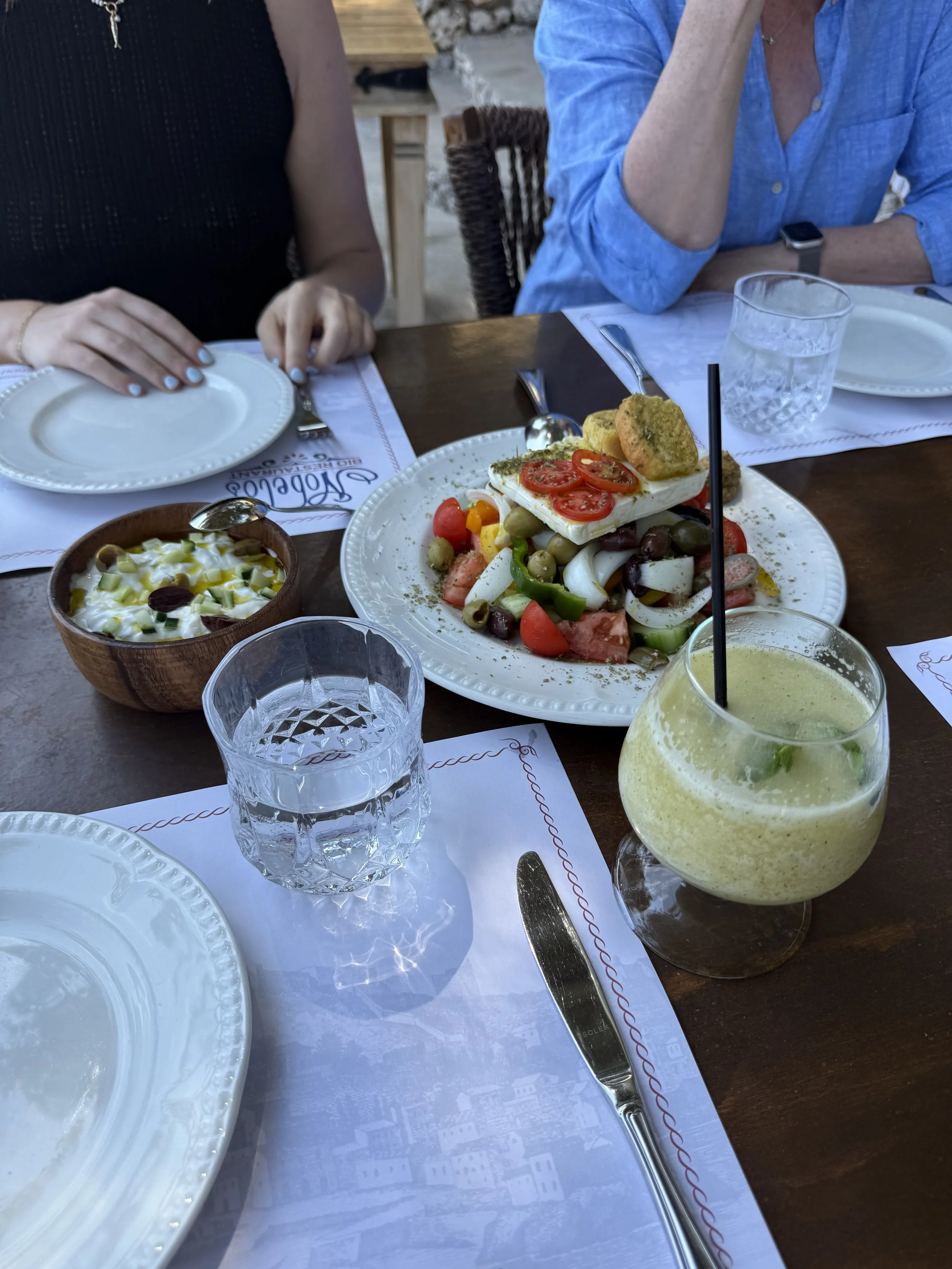  Classic Greek salad with tomatoes, feta, and olives on a restaurant table with a green cocktail in view. 
