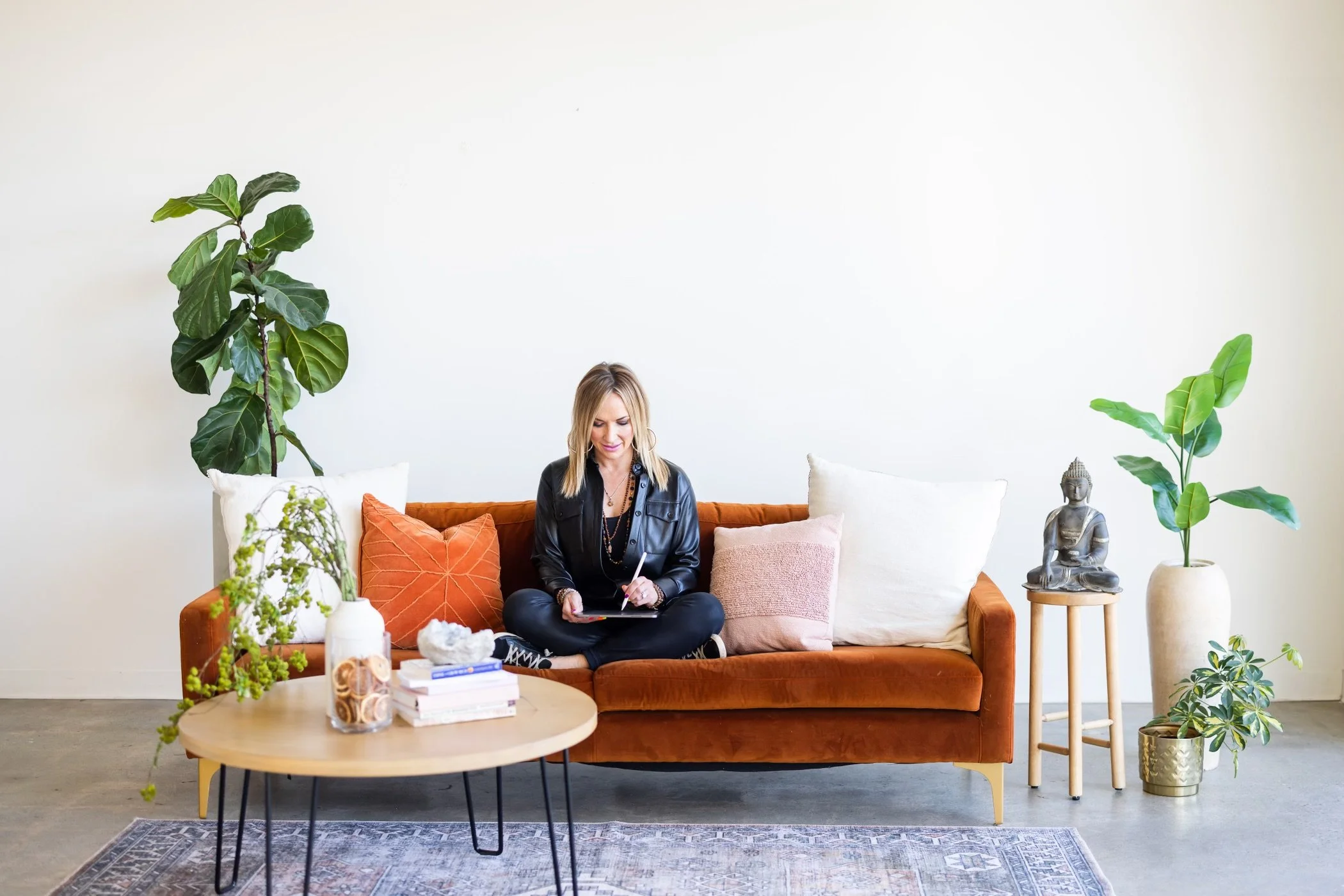 A woman with blonde hair sitting on an orange sofa, writing in a notebook, surrounded by plants and decorative pillows with a minimalist white wall background.