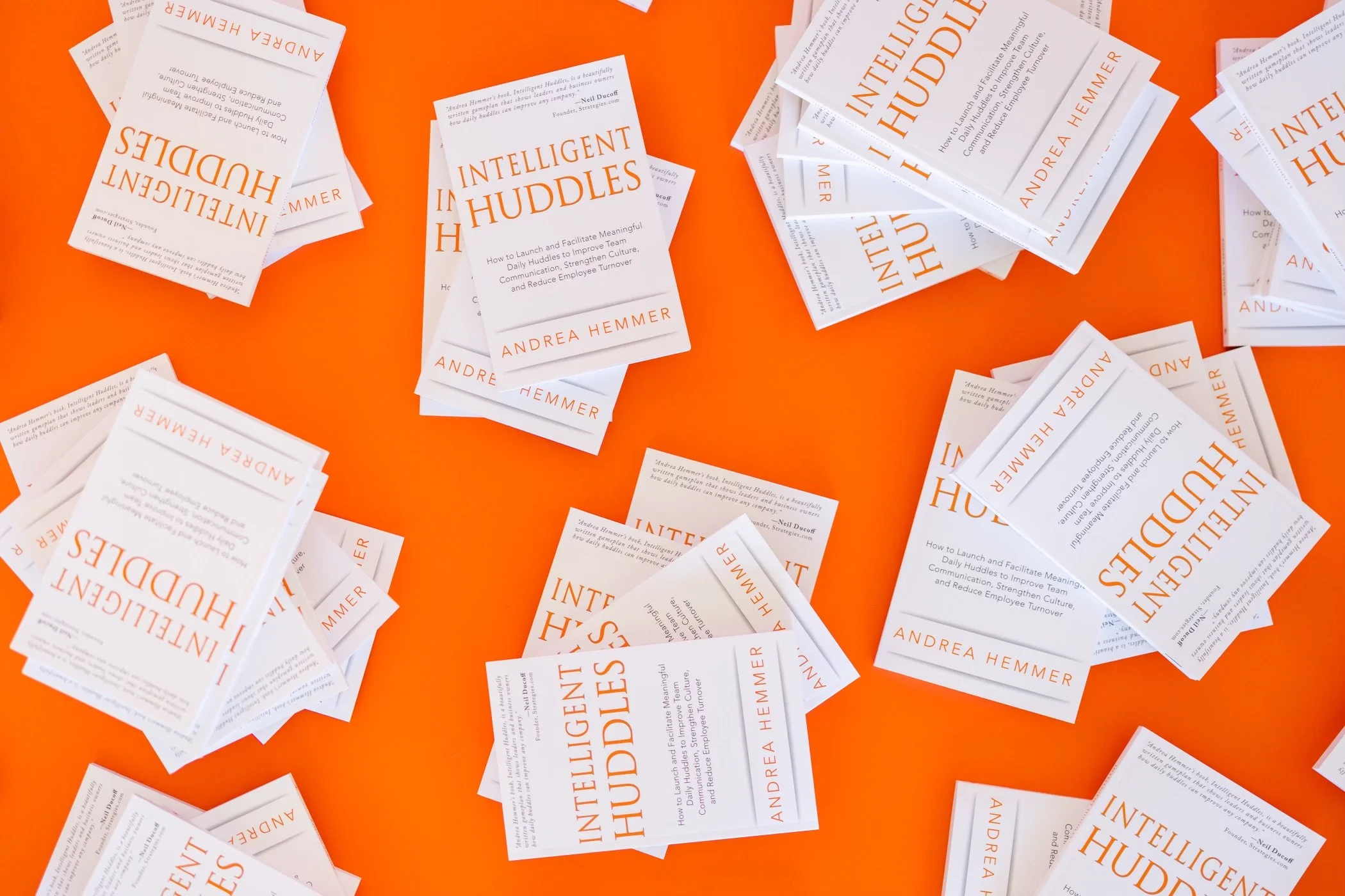Several piles of the Intelligent Huddles book, written by Drea Hemmer, on a bright orange backdrop.