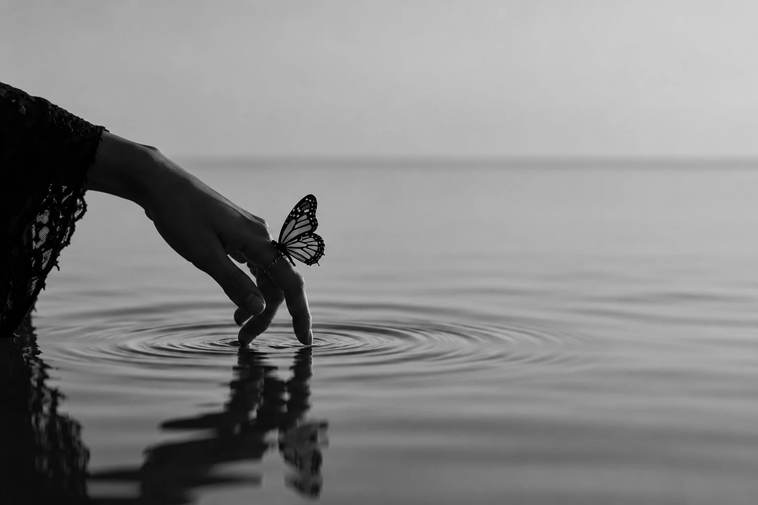 Svartvit fotografisk bild av en hand som vidrör vatten och skapar ringar på ytan, med en fjäril på fingret. Bilden symboliserar frigörelse, läkning och personlig utveckling efter narcissistiskt våld och uppväxt i ett narcissistiskt familjesystem.