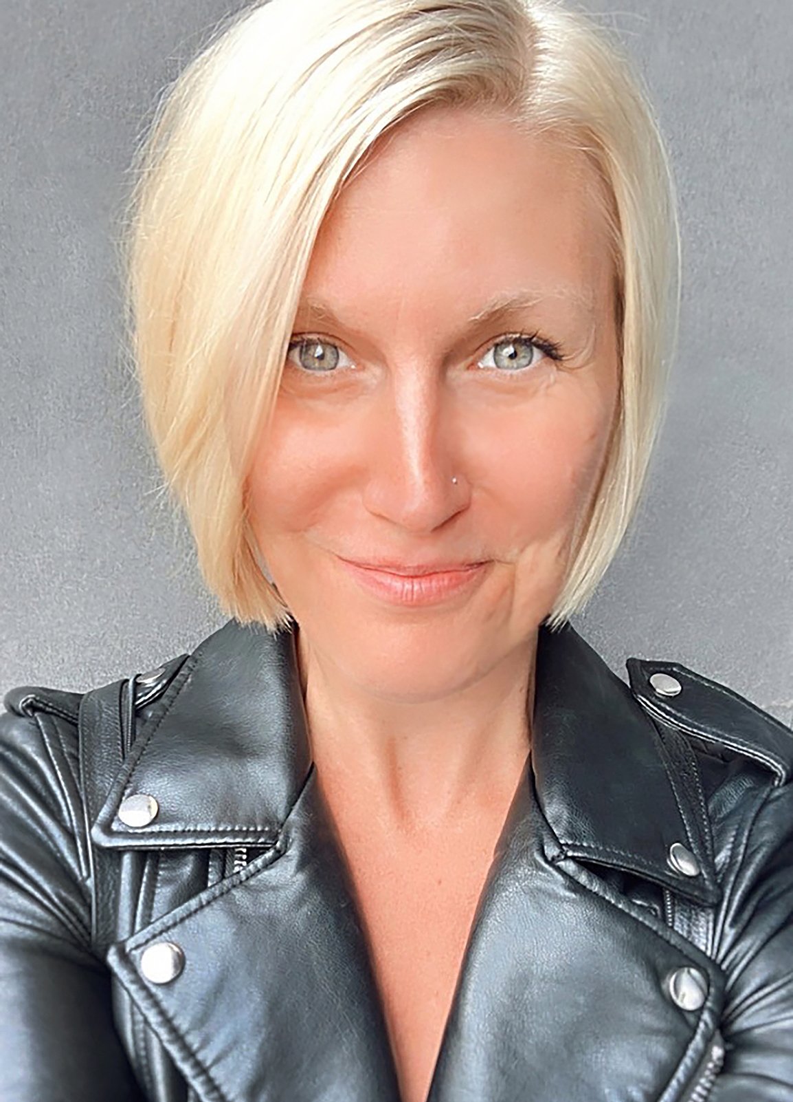 A woman with blonde hair and blue eyes taking a selfie. She is wearing a black leather jacket and smiling softly. The background is a plain gray wall.