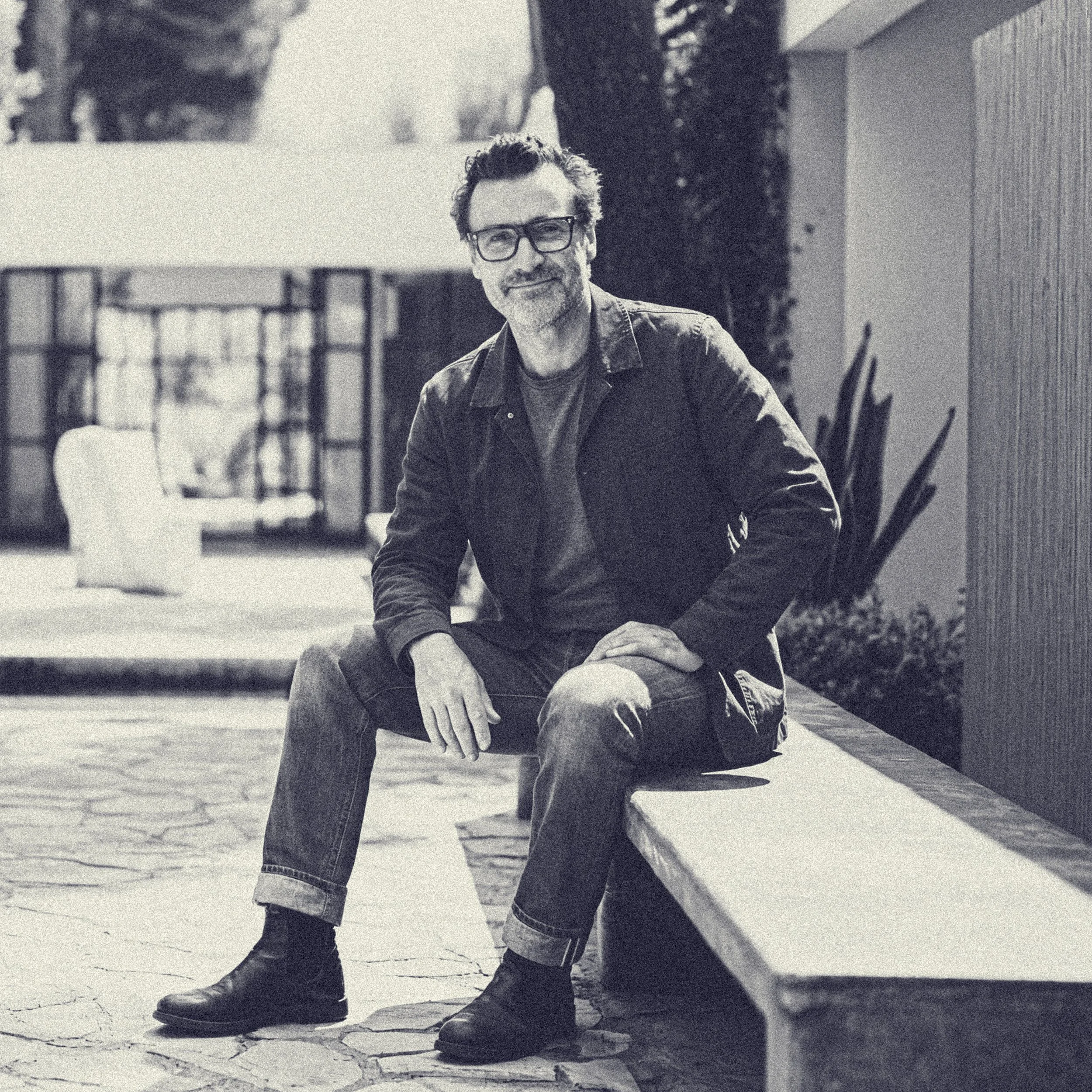 A man with glasses and a beard relaxing outdoors, sitting on a concrete sidewalk next to a wood-paneled building, with greenery and trees in the background.