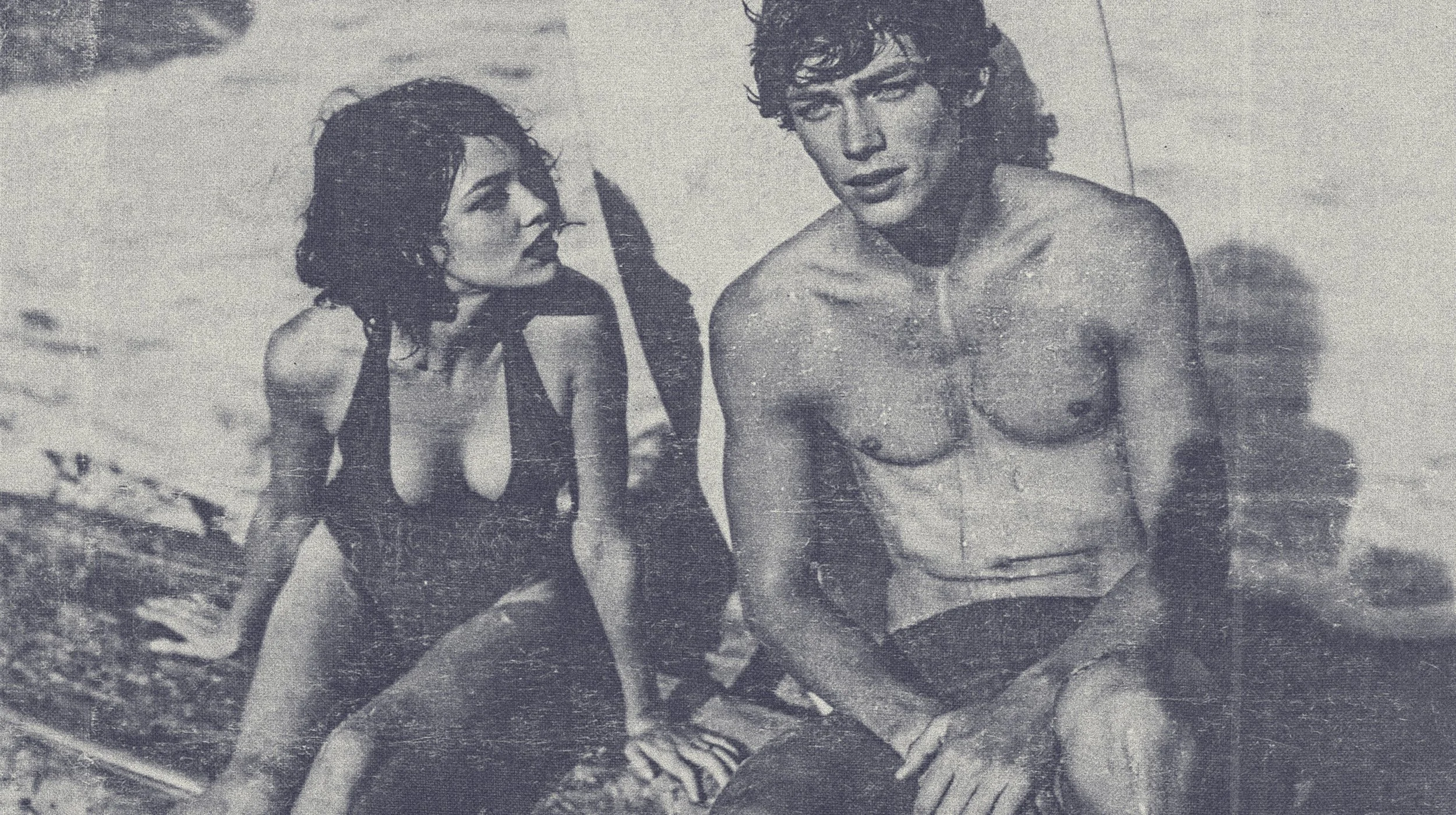 A black and white photo of a woman and a shirtless man with messy hair, sitting outdoors near water. The woman is looking at the man, who is facing forward, with the woman slightly to his left. They are on a sandy or rocky surface.