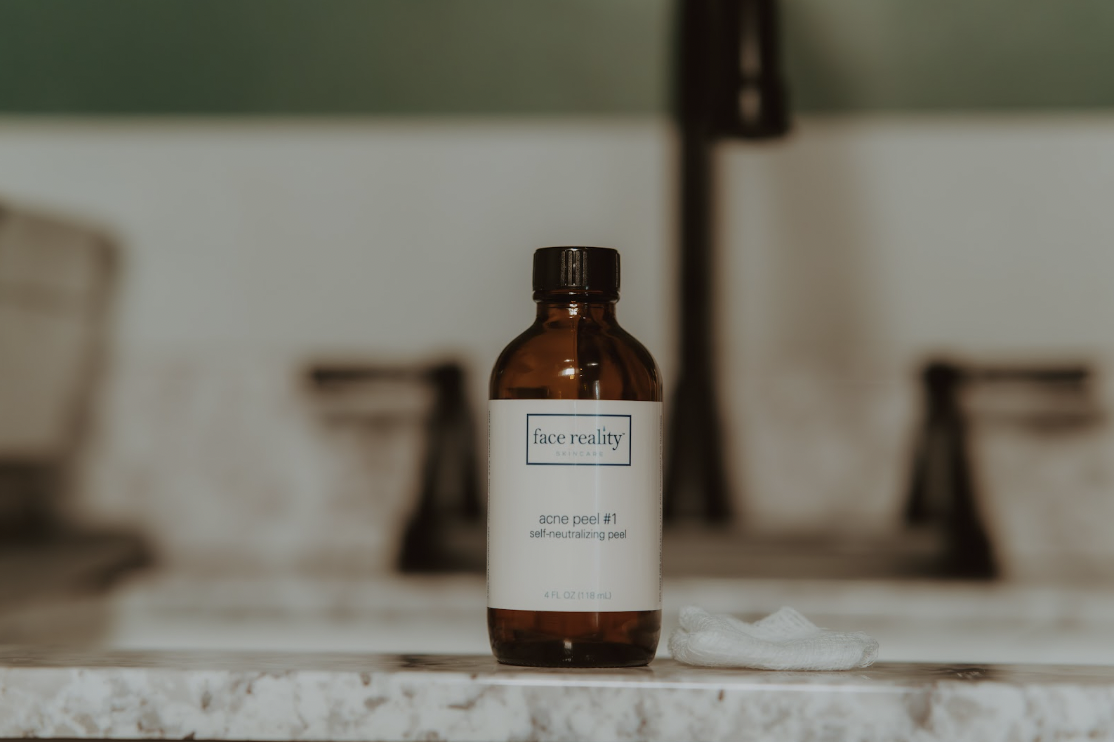 A small amber glass bottle labeled 'face reality acne peel #1 self-neutralizing peel' on a bathroom countertop with a white cloth next to it.