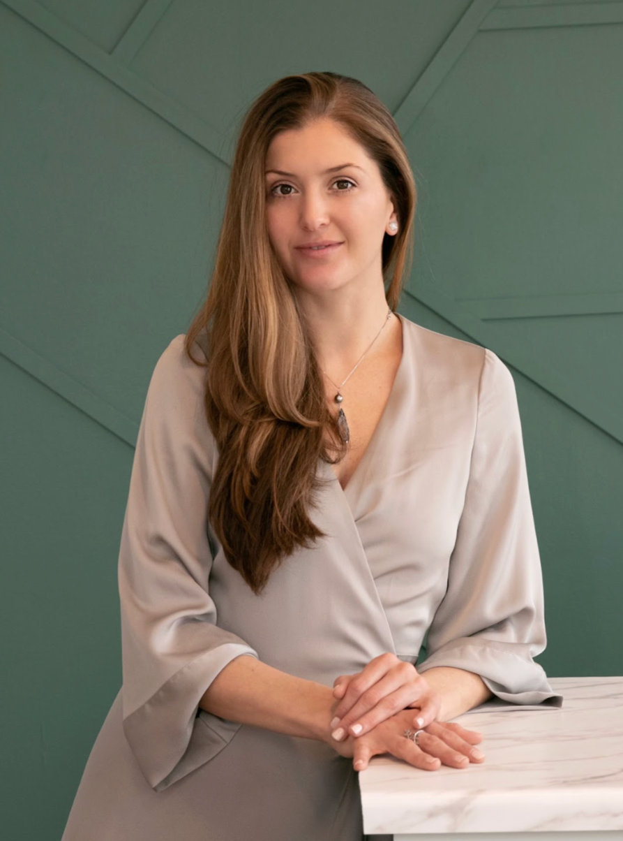 A woman with long brown hair sitting on a yellow armchair near windows, wearing a light-colored blouse, and smiling while resting her chin on her hand.
