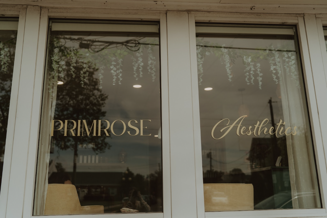 A storefront window with the words "PRIMROSE" in serif font on the left and "Aesthetics" in cursive on the right, reflecting outdoor scenery with trees and power lines.