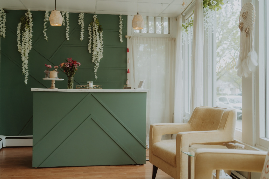 A cozy interior of a cafe or boutique with a green reception counter decorated with flowers, hanging white floral garlands, beige armchairs, and large windows with white curtains letting in natural light.