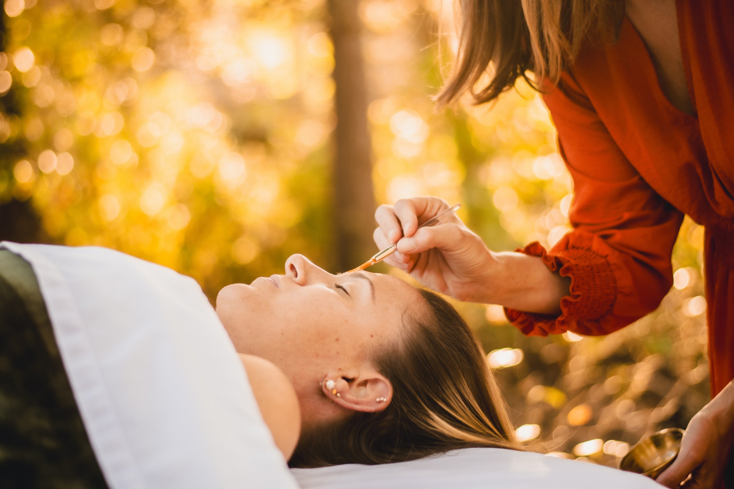 A woman lying outdoors with her eyes closed, while another woman applies makeup with a brush during sunset or golden hour.
