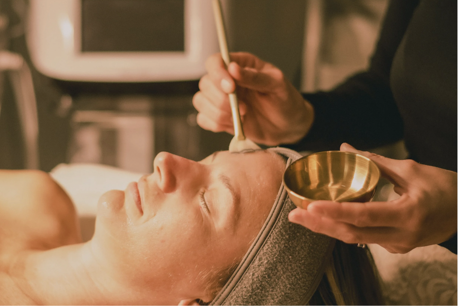 A woman lying on a massage table receiving a facial treatment from a practitioner who is holding a bowl in one hand and applying a substance on her face with a brush.