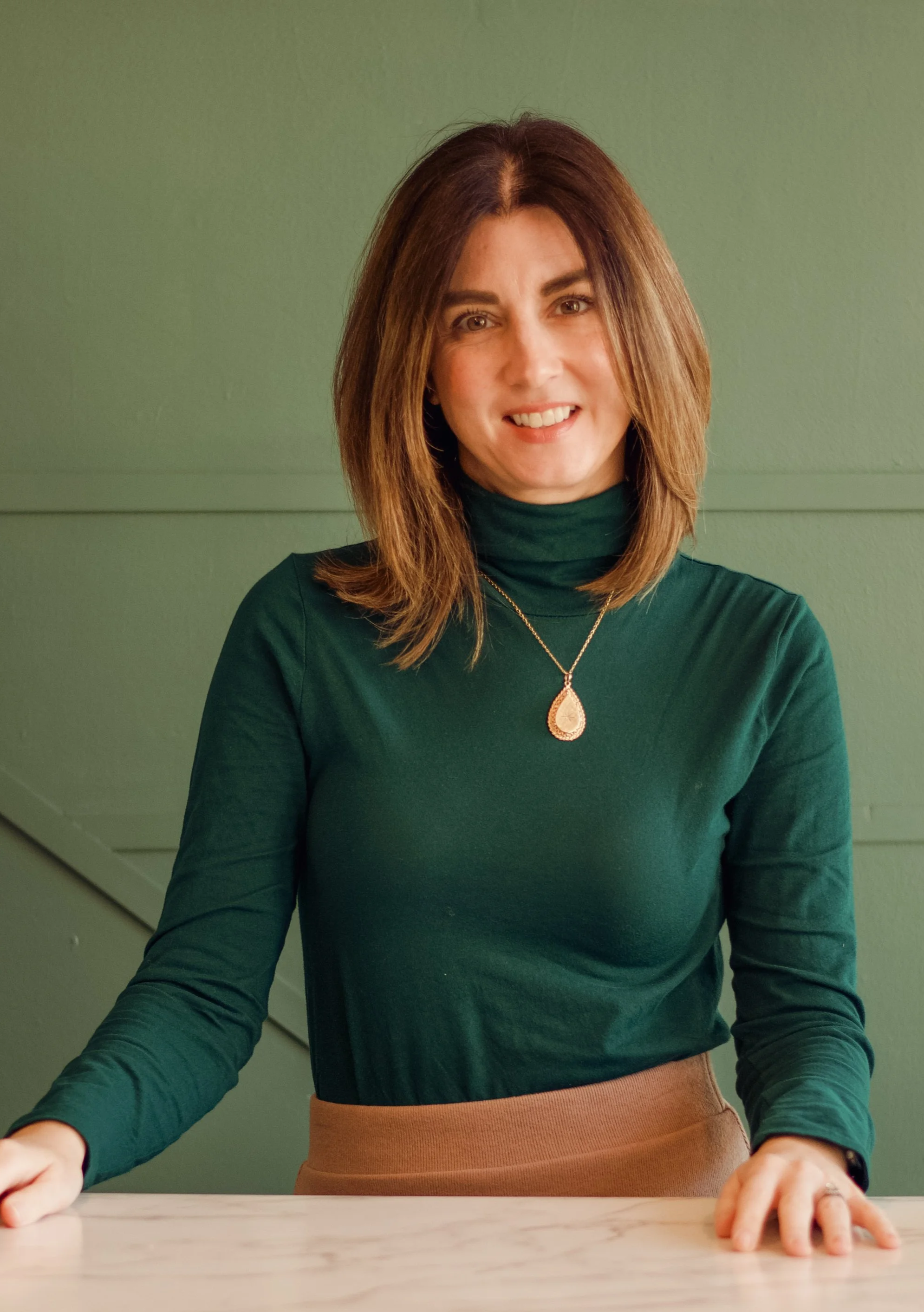 A woman with shoulder-length brown hair, wearing a black shirt and a gold necklace, standing and smiling in front of a plain light-colored wall. She is wearing a checkered jacket in beige and black.