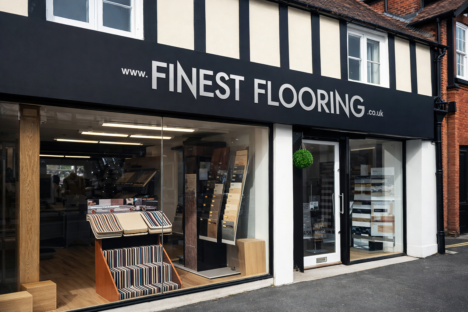 Exterior view of a flooring store called 'Finest Flooring' with large glass windows displaying sample flooring options and carpets, and a black sign with the store's website.