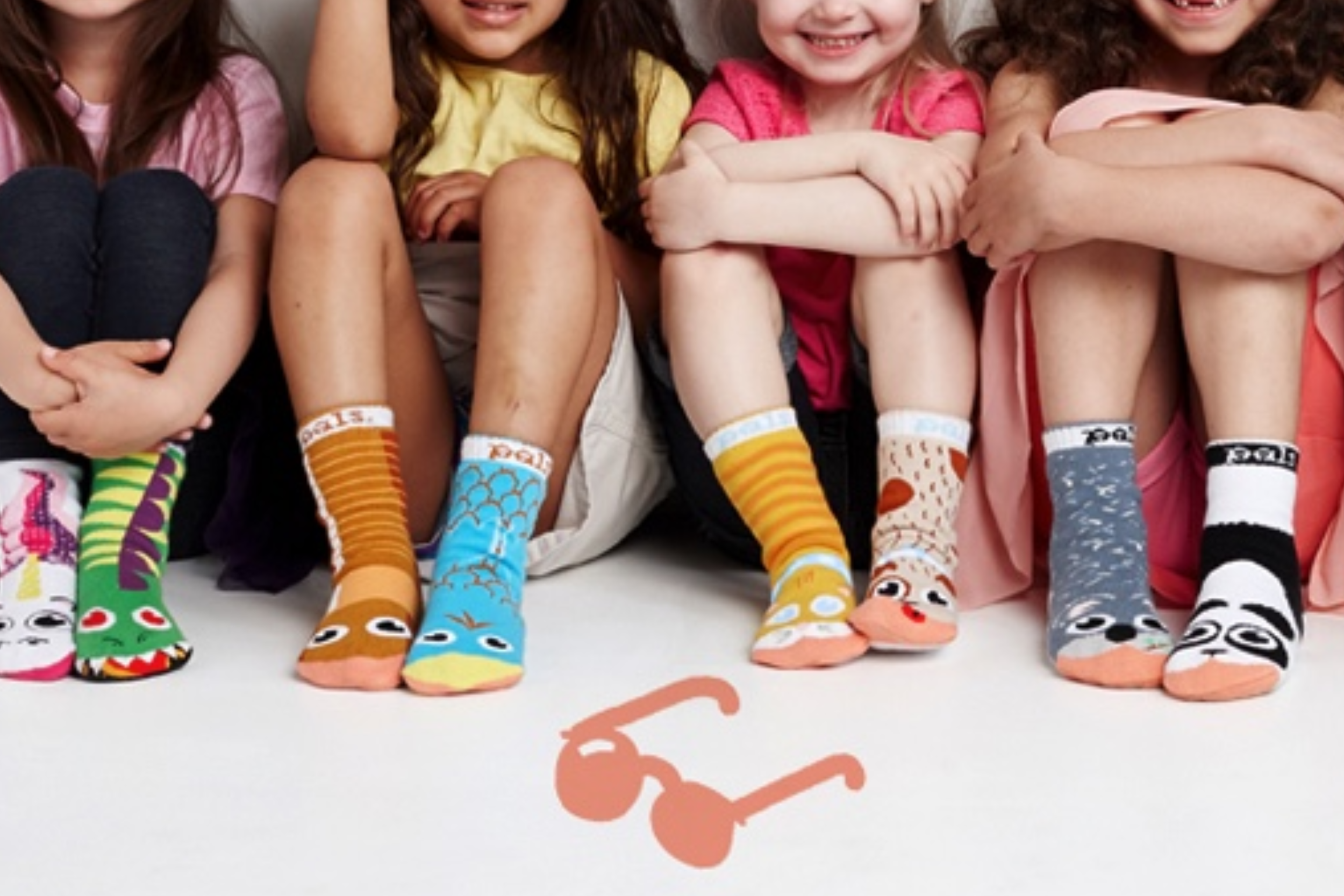 Six children sitting on the floor with their legs extended, wearing colorful socks with playful designs. There is a small illustration of sunglasses on the floor in front of them.