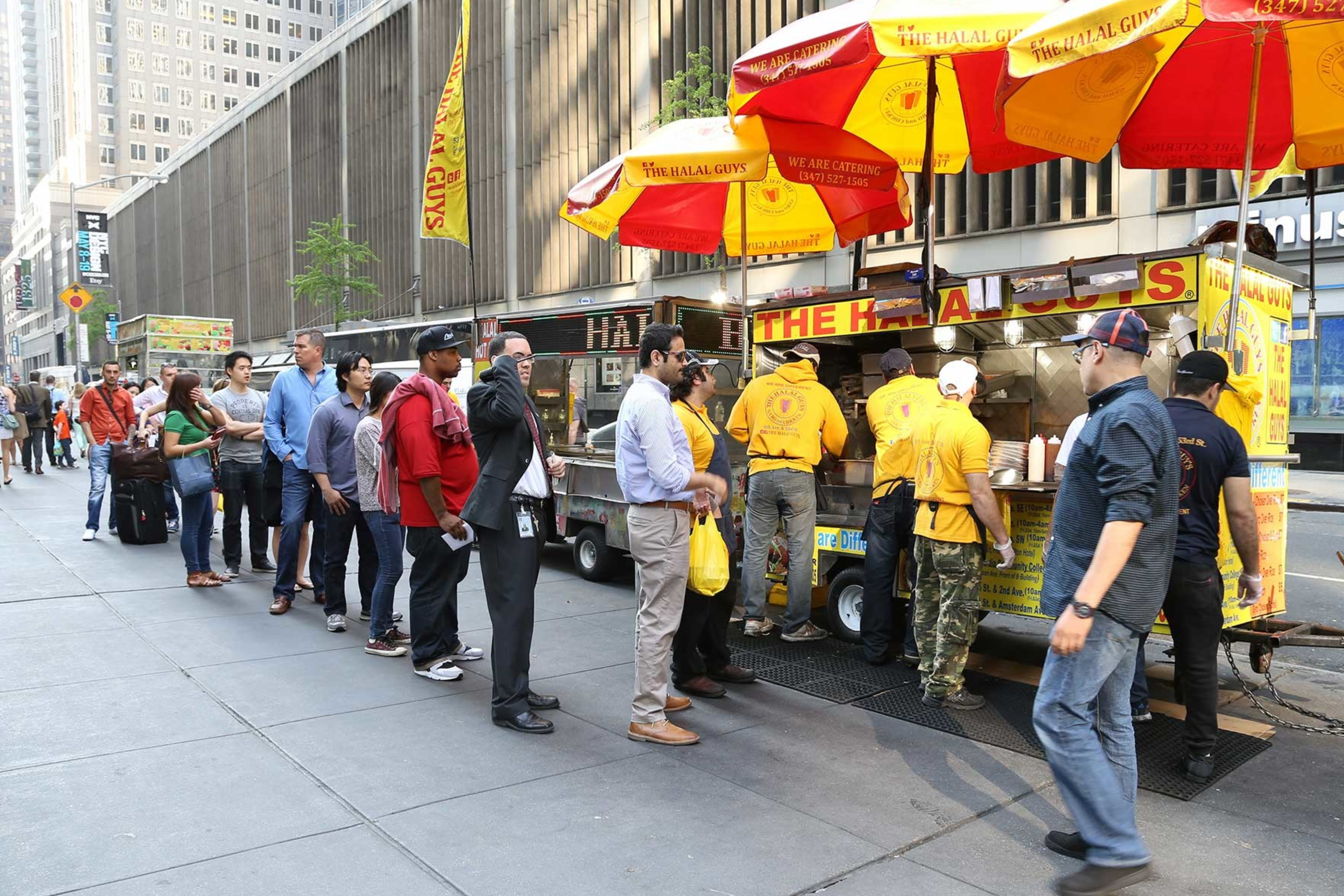Line of people waiting to order food from a yellow and red food truck on a city street.