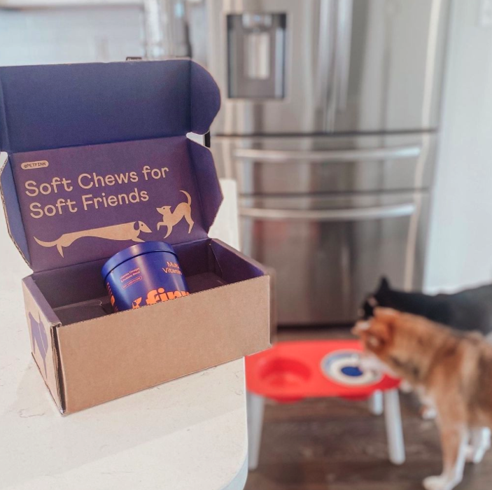 Open cardboard box labeled "Soft Chews for Soft Friends" with a container inside, placed on a kitchen counter. Two dogs are in the background near a red and blue dog bowl.