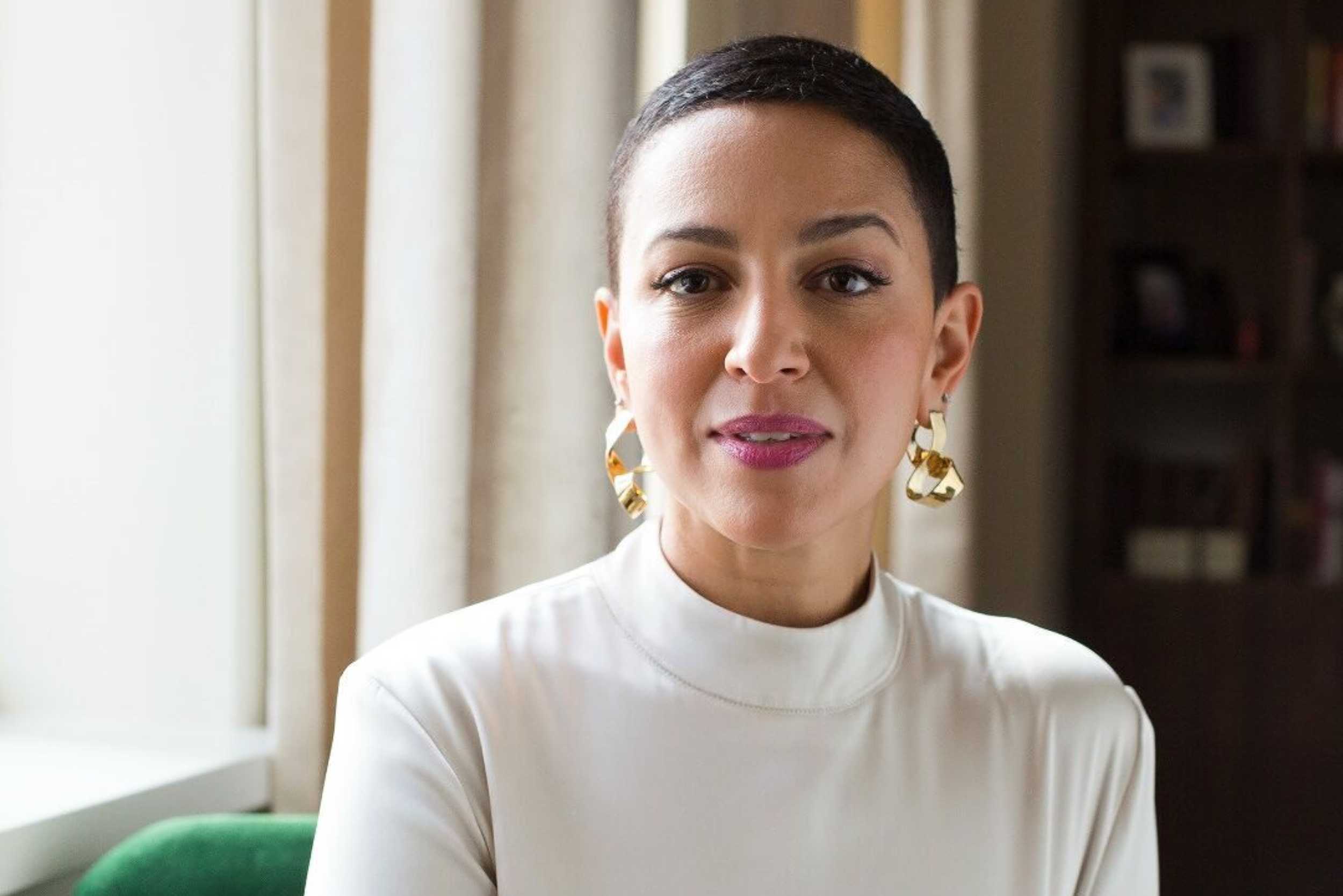 A woman with short dark hair, wearing a white high-neck top and large gold earrings, sitting indoors near a window with curtains.