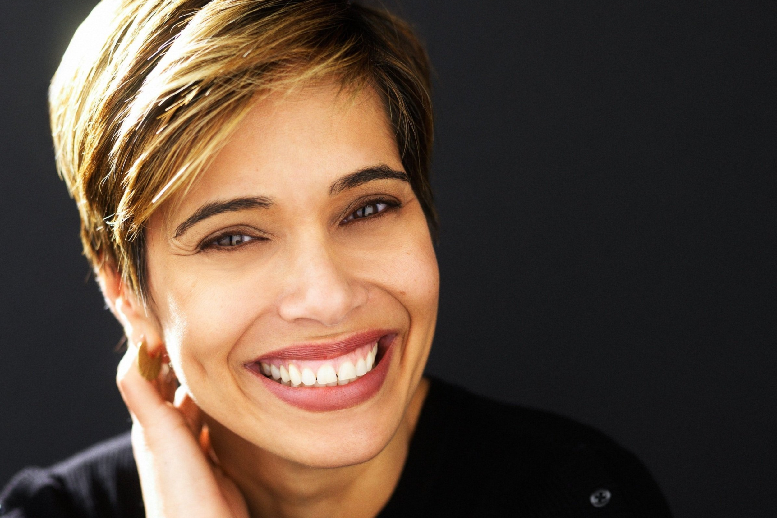 Close-up of a smiling woman with short, light brown hair, wearing a black top, against a dark background.