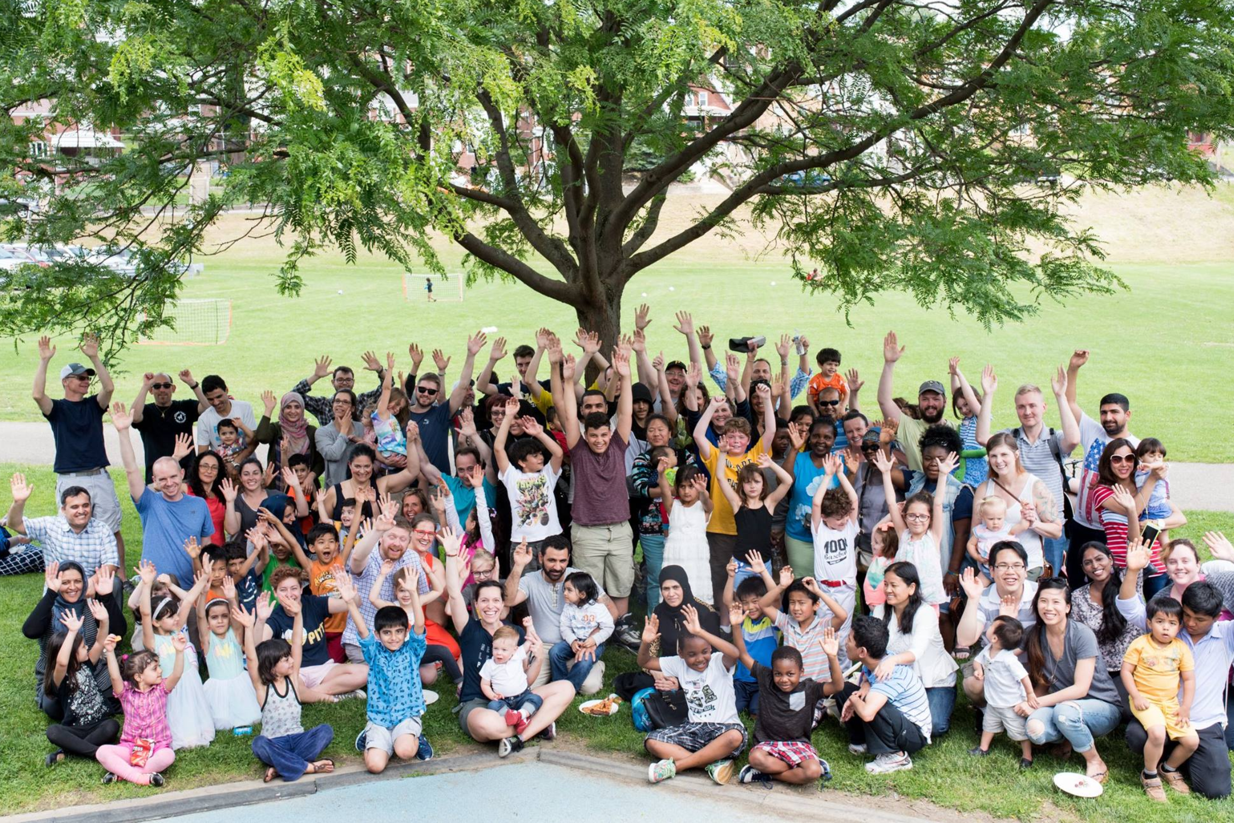 Large diverse group of people gathering under a tree in a park, smiling and waving at the camera, with children in the front and a grassy field in the background.